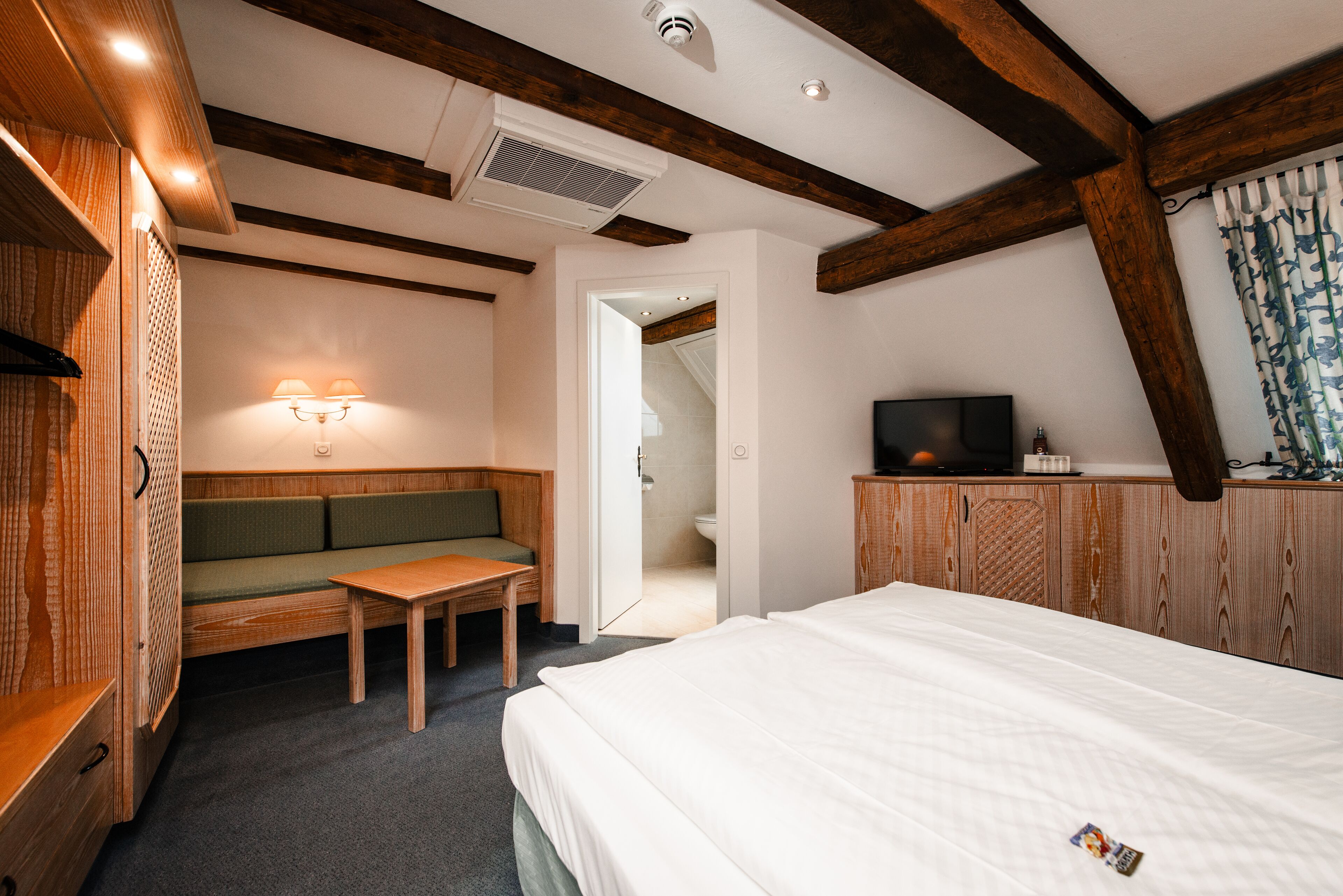 Une chambre d’hôtel avec un banc en bois, un lit, une télévision et une salle de bain visible à travers une porte ouverte.
