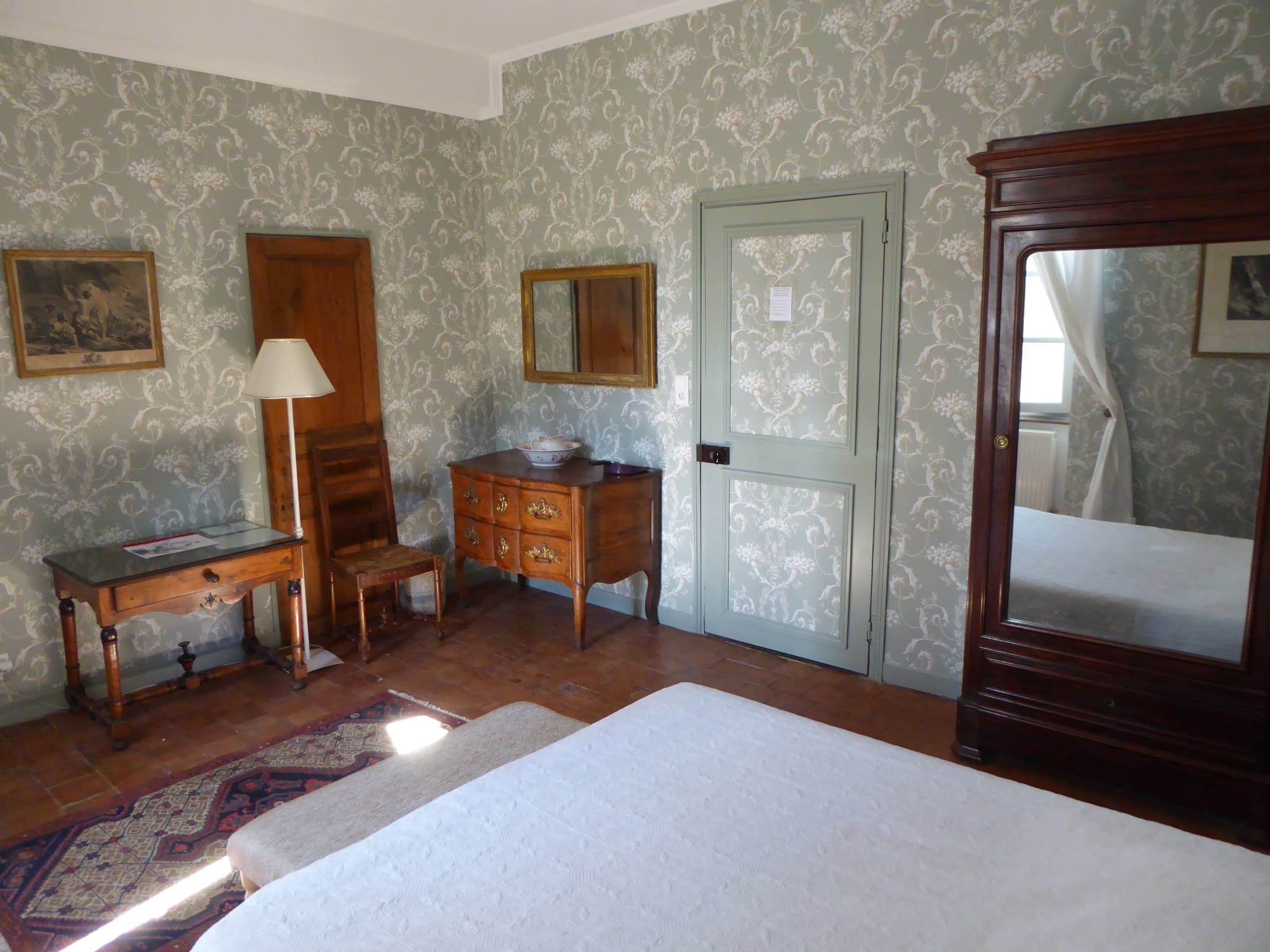Habitació amb llit, una petita ampolla de fusta, una cadira, escriptori amb làmpada i porta amb mirall.