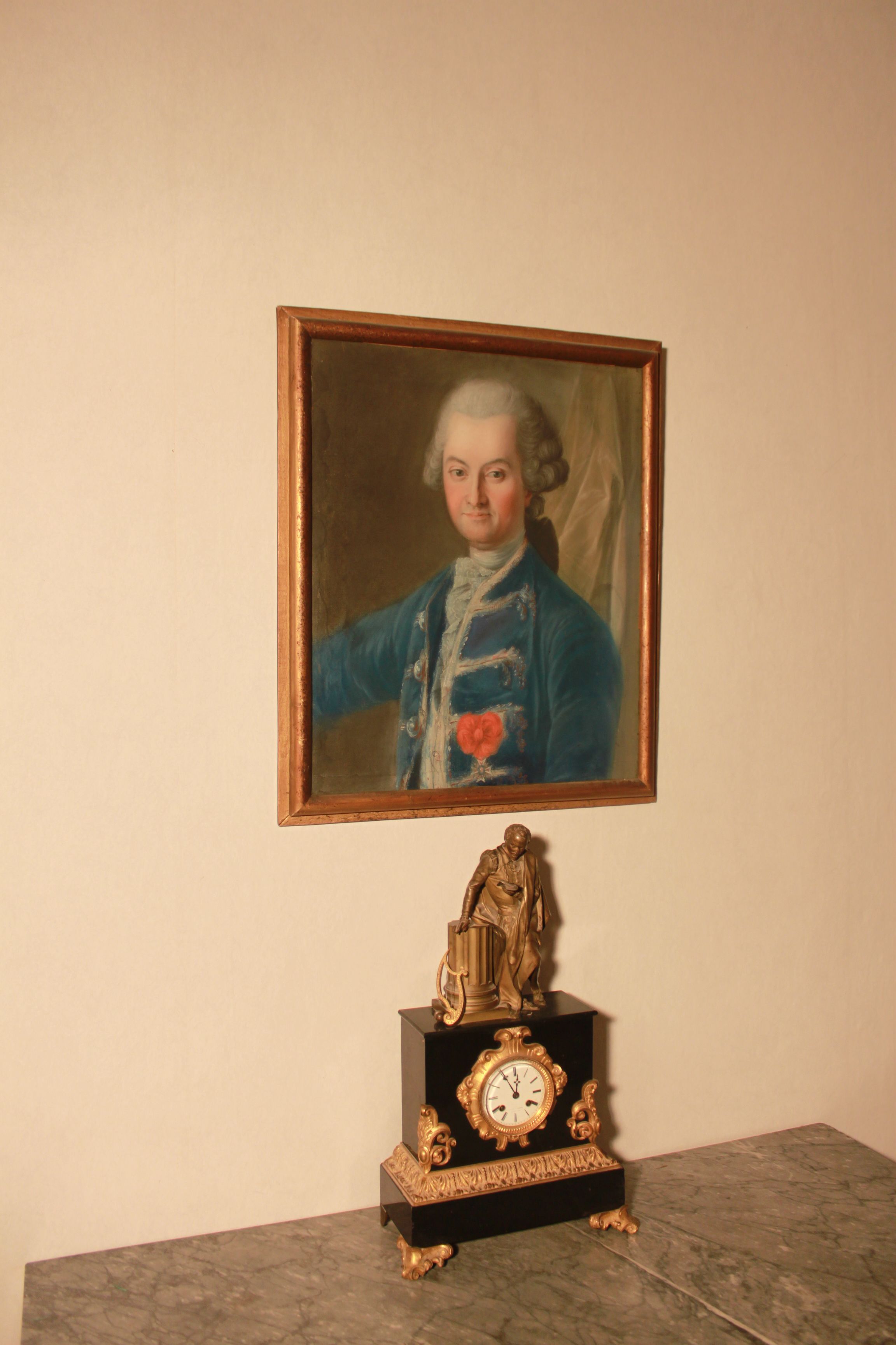 Un retrato enmarco de una persona con vestimenta histórica, una estatua y un reloj sobre una mesa de mármol.