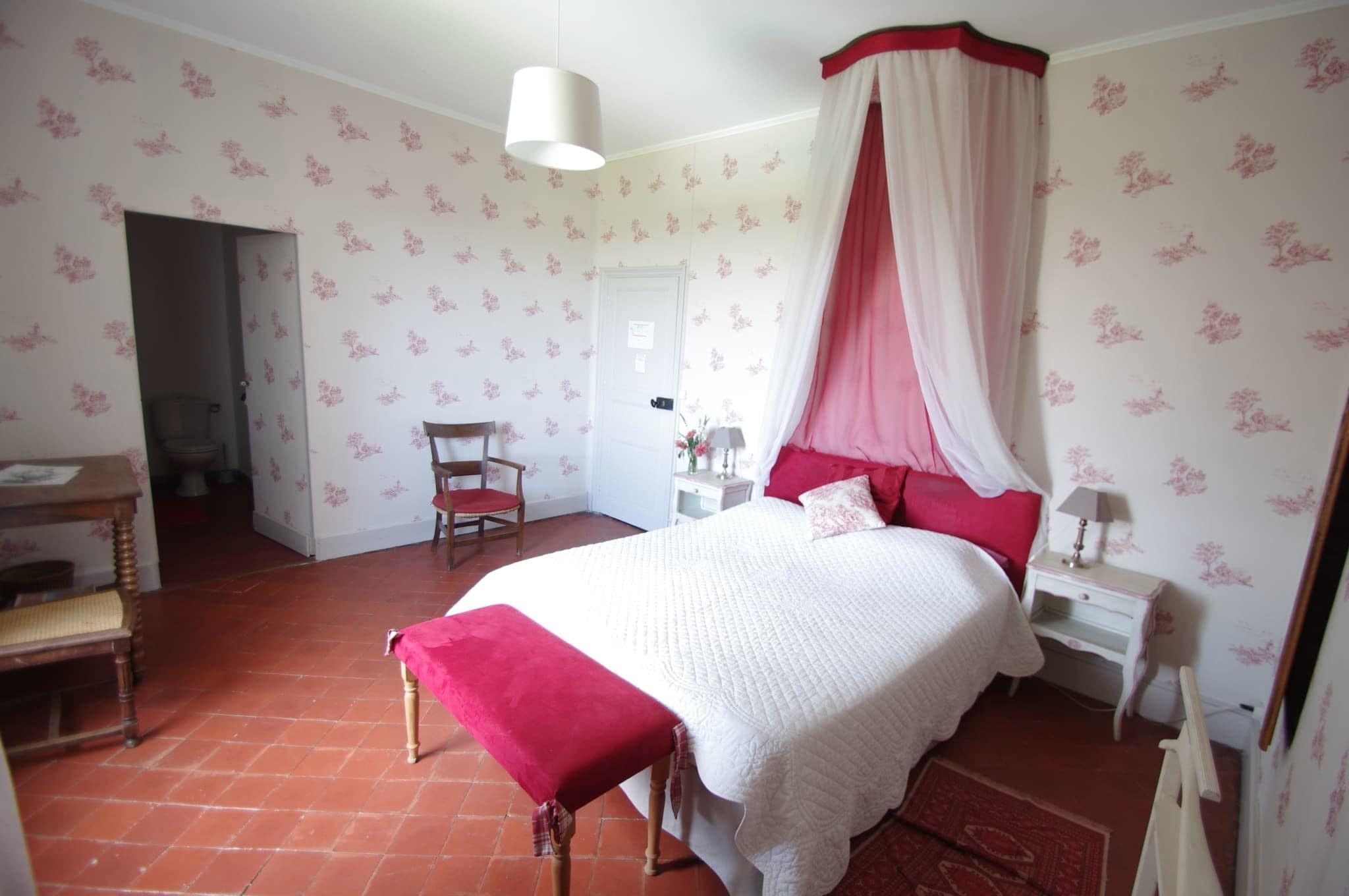 A bedroom with a four-poster bed, a bedside table, a chair, and a red bench.