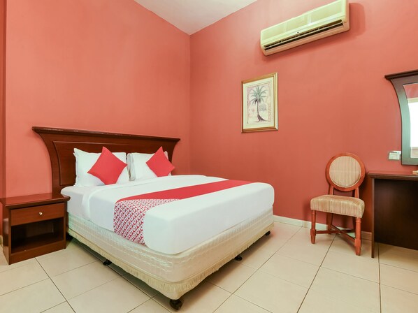 A hotel room with a large bed, a nightstand, a chair, a desk with a mirror, and an air conditioning unit.
