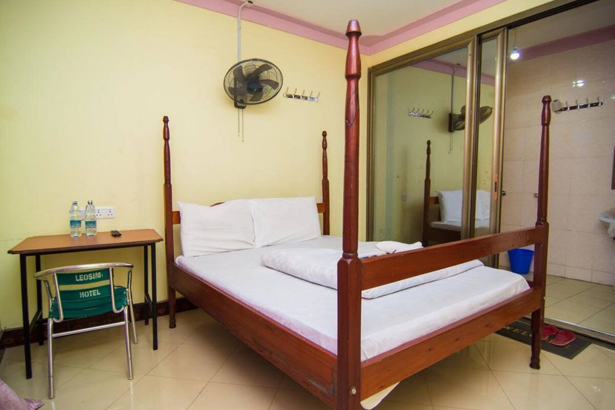 A four-poster bed with white linens, a wooden nightstand with a green chair, and a mirror reflecting the room.