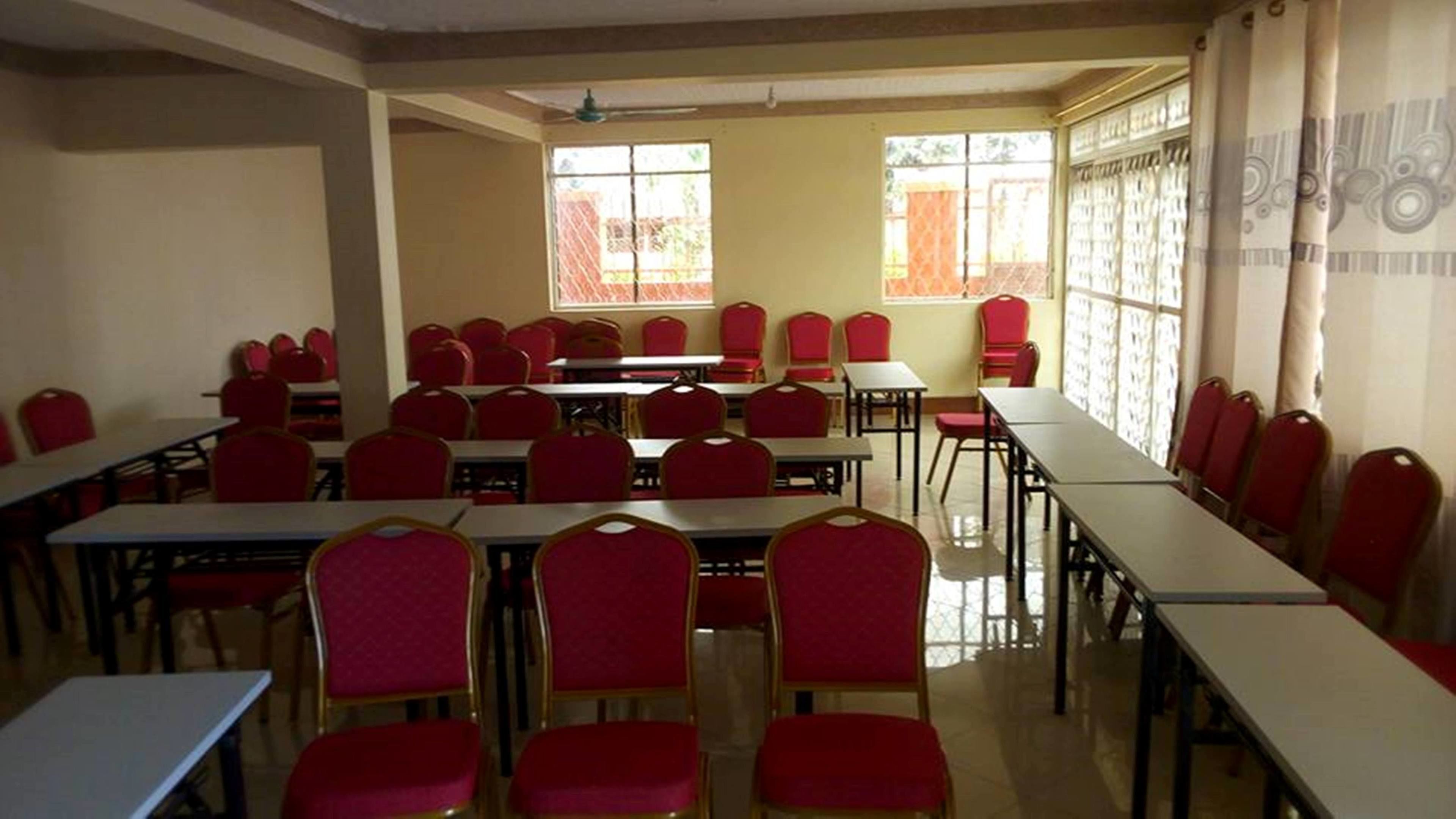 Une salle de classe avec des rangées de bureaux et des chaises rouges, de grandes fenêtres et un ventilateur au plafond.