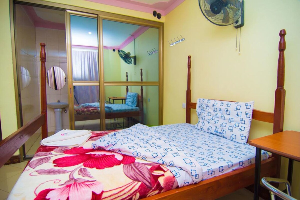 A bedroom with a bed, a wooden nightstand, a chair, a small table, a fan, and a sliding glass door leading to a bathroom.