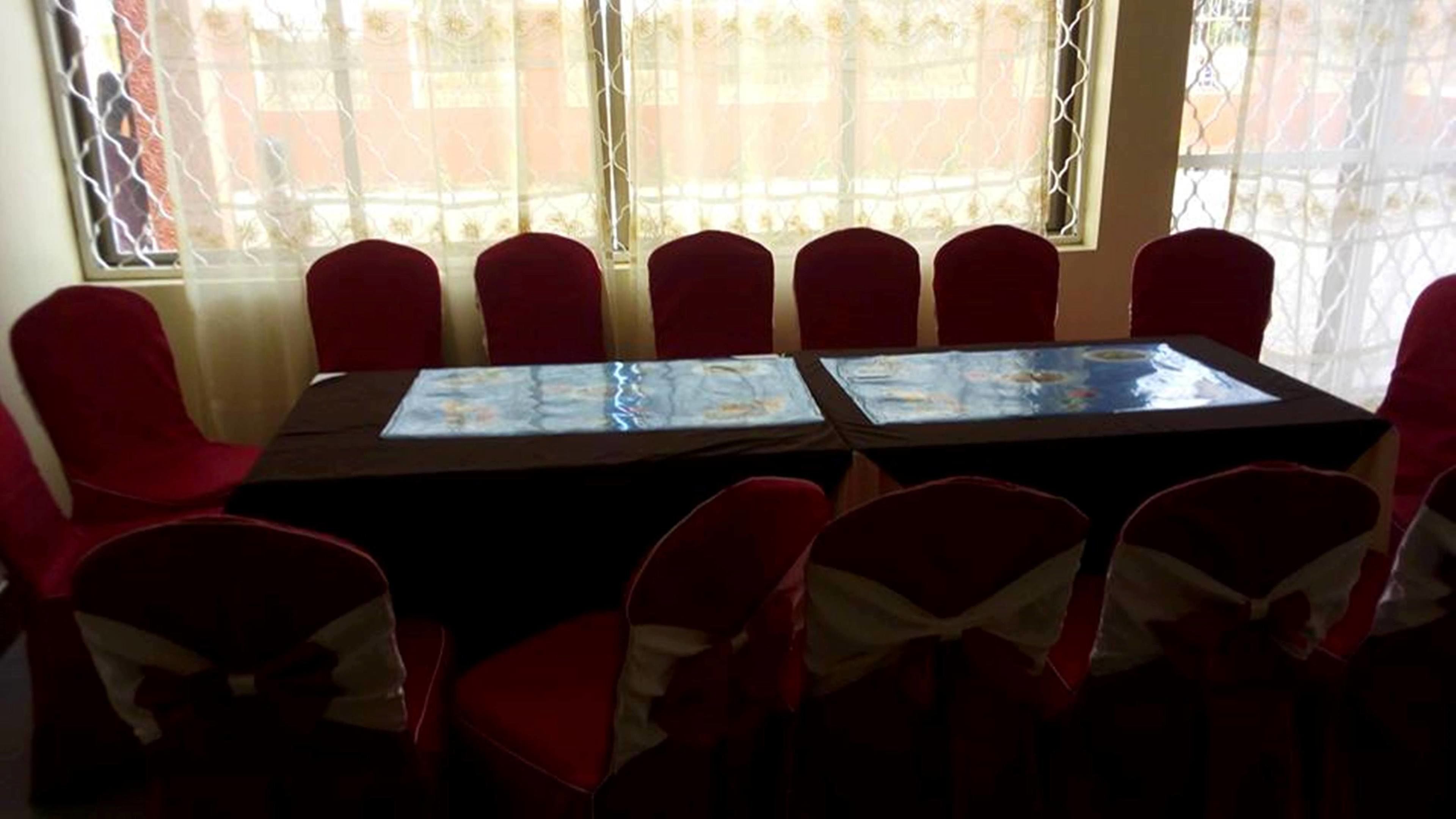 A conference room with a long table and red chairs, large windows with decorative curtains, and a view of buildings outside.