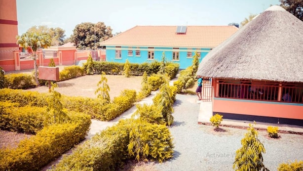 A courtyard with a thatched-roof building, a blue building with multiple windows, and well-maintained greenery. - Leosim Hotel (Pakwach)