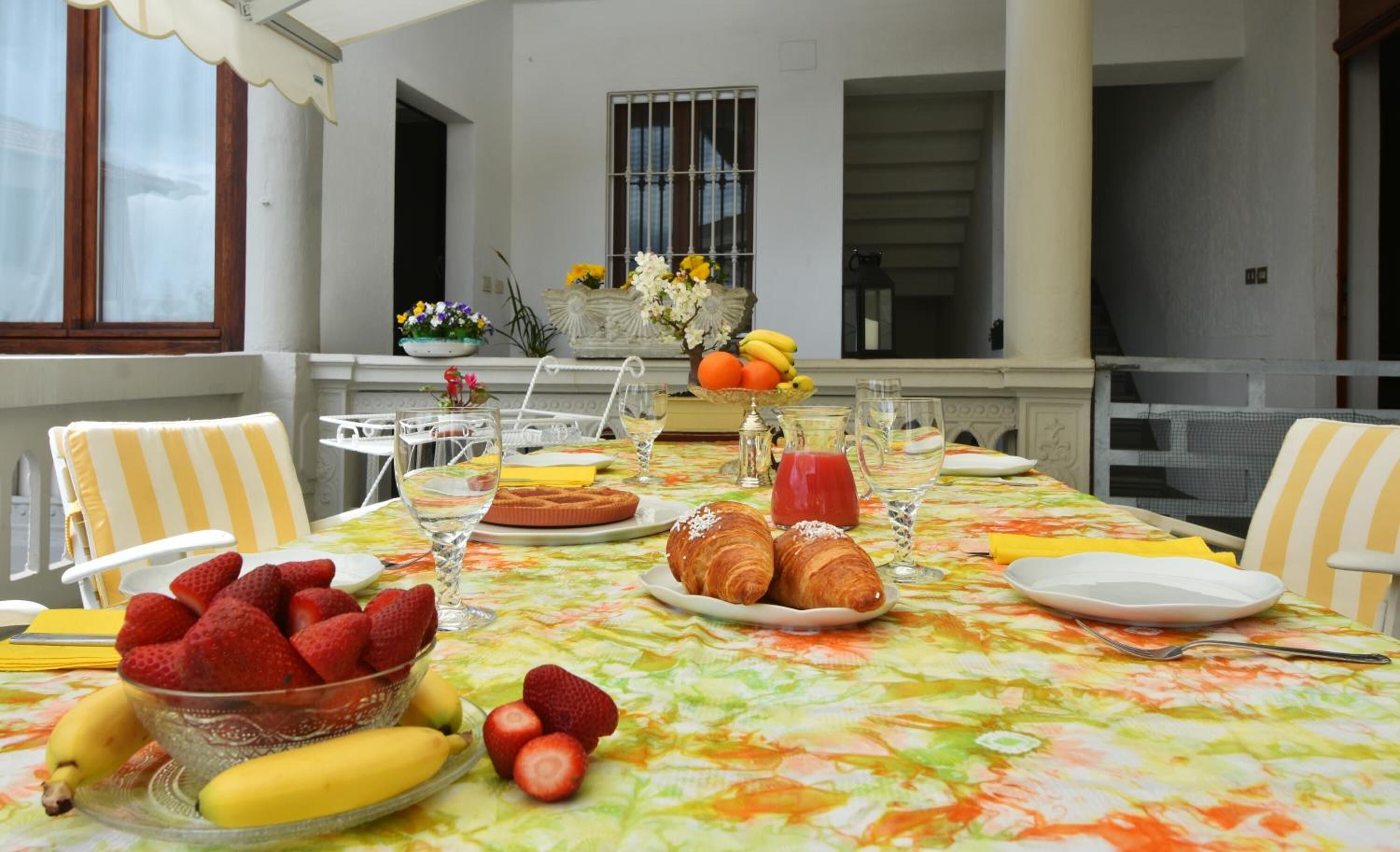Una mesa de comedor dispuesta con frutas, pasteles y bebidas, con una ventana y flores decorativas al fondo.