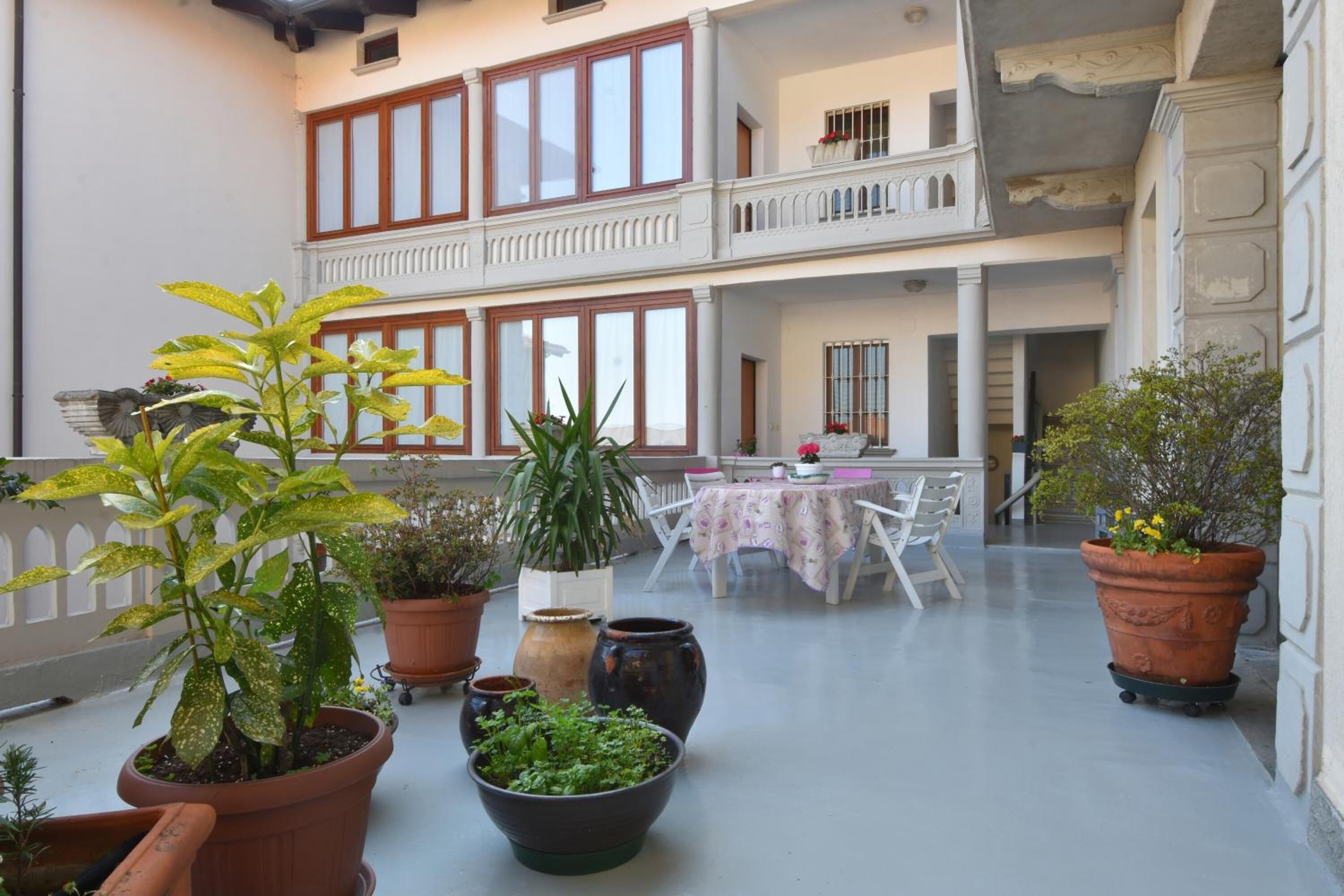 A spacious balcony with multiple potted plants, a dining table with chairs, and a view of the building's interior.