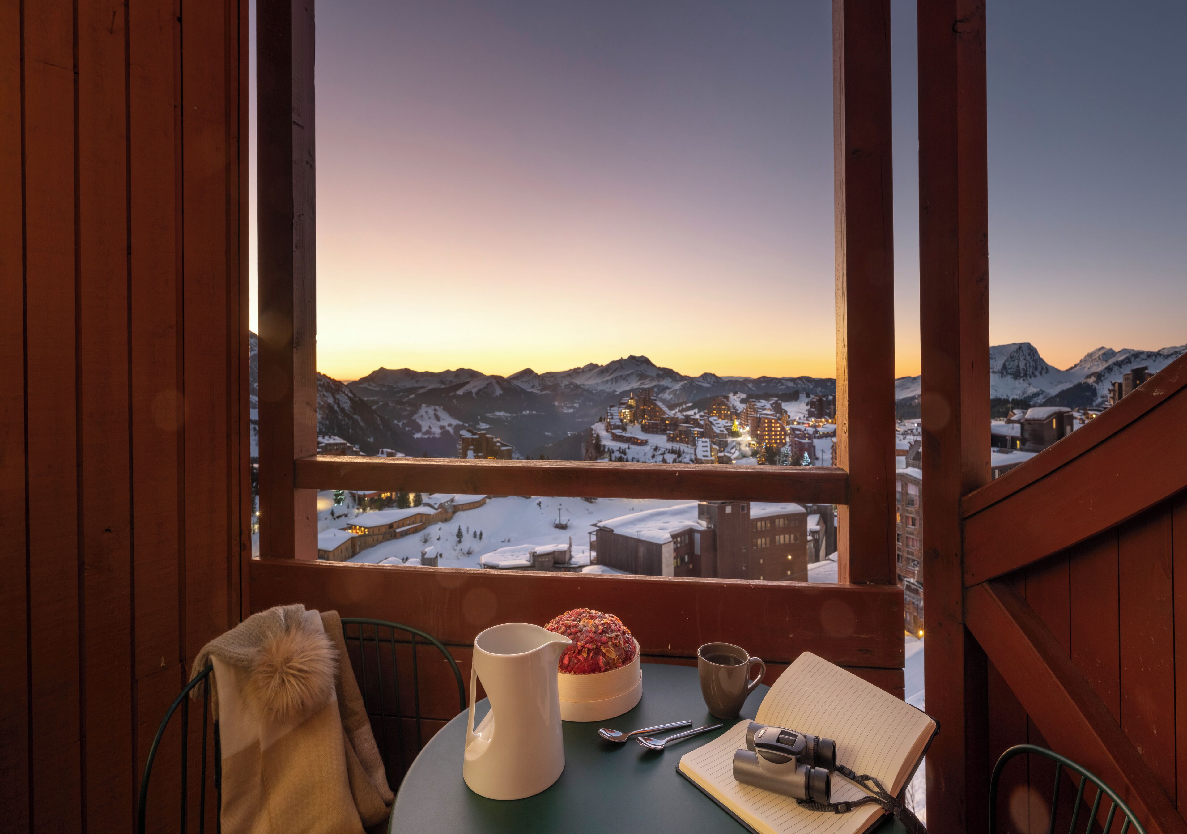 A wooden cabin with a table set for breakfast, a view of snow-covered mountains, and a sunset.