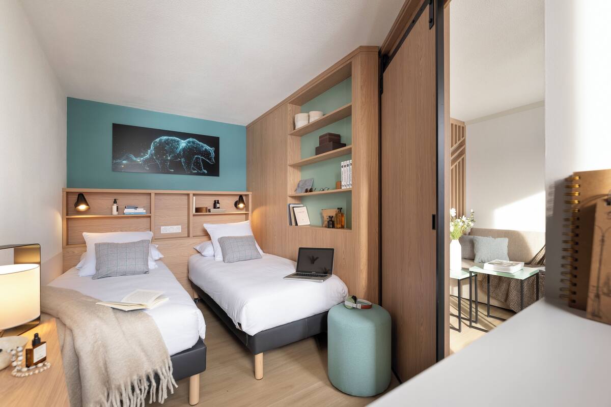 A modern bedroom with two beds, a laptop on one bed, a desk with a chair, and a large wall art piece.