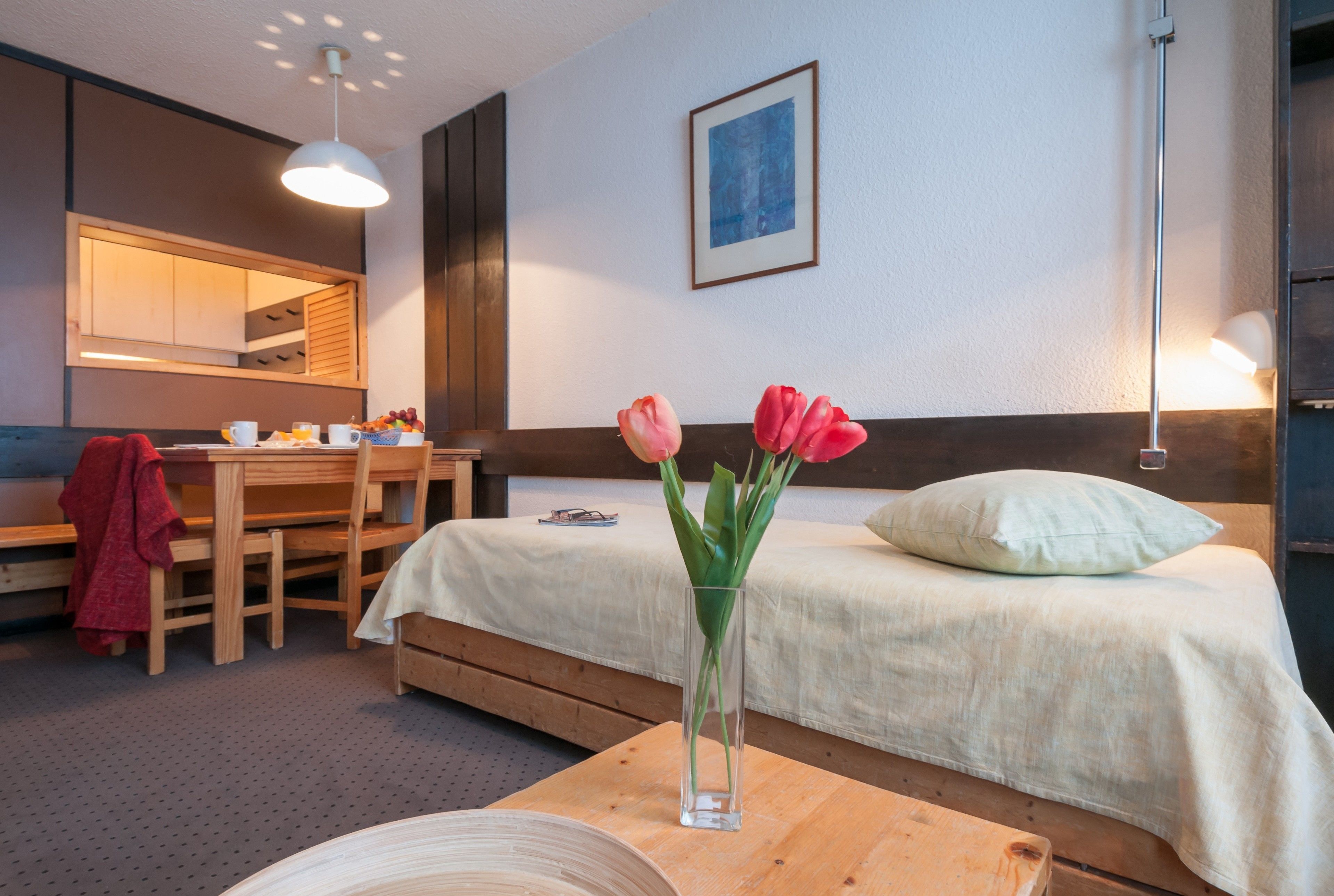 A hotel room with a bed, a desk with a chair, a vase with flowers, a lamp, and a framed picture on the wall.