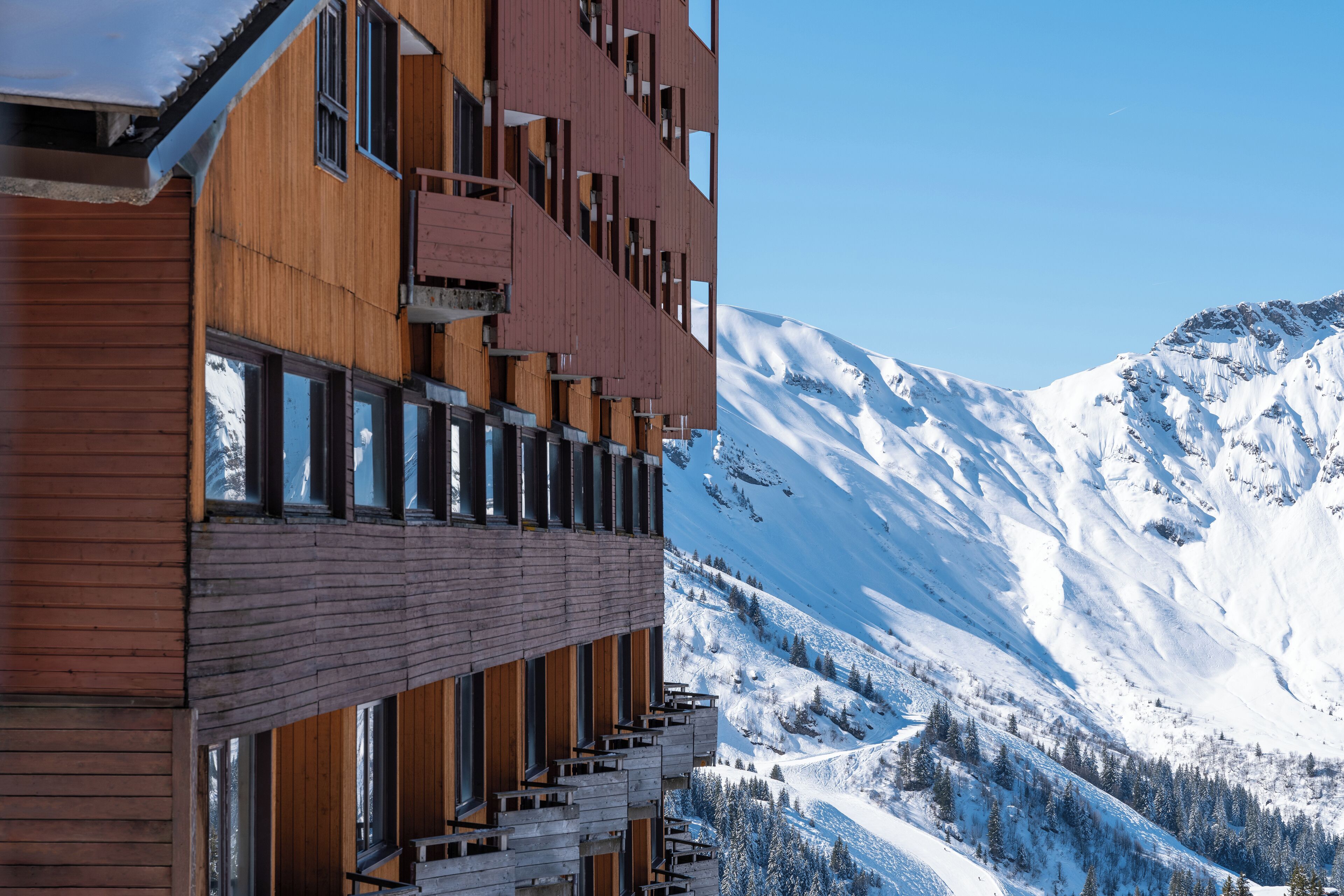 A modern building with wooden exterior and large windows, overlooking a snow-covered mountain.