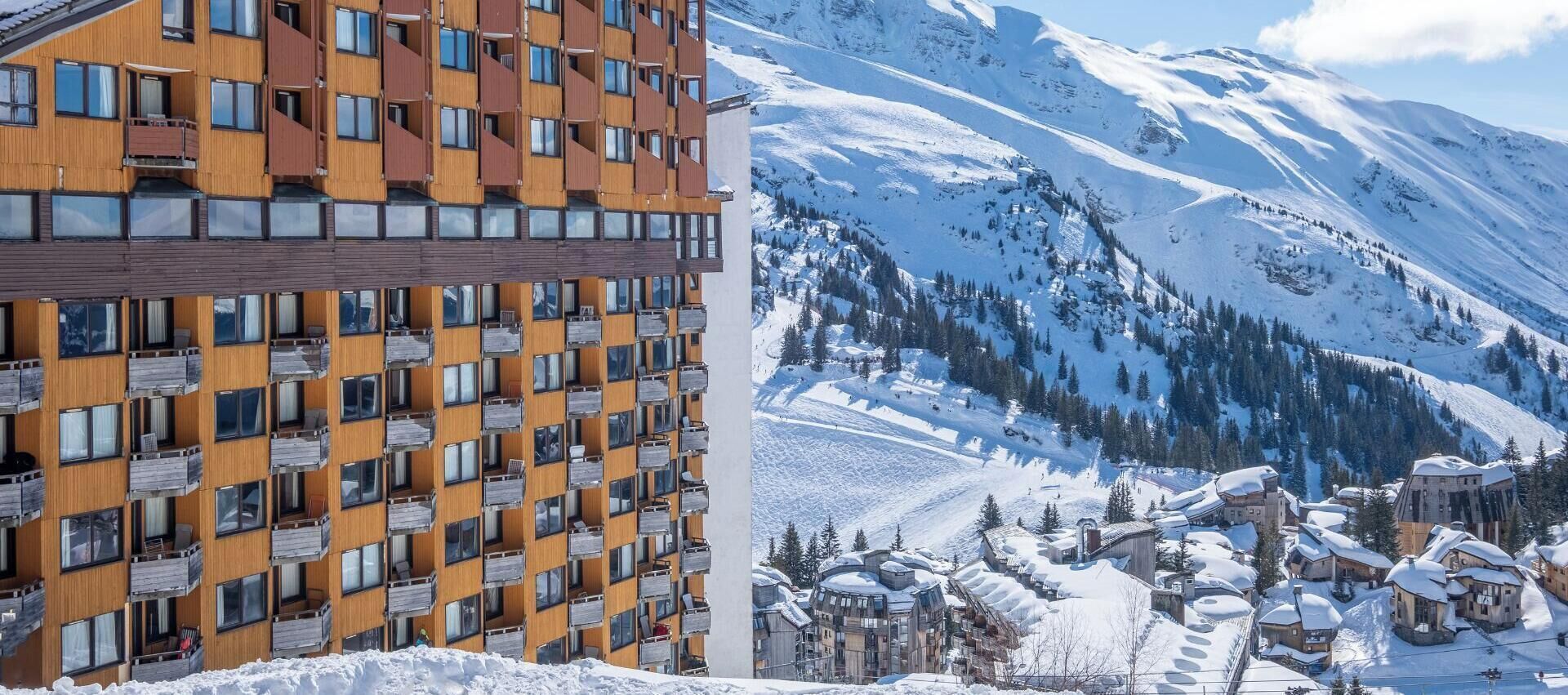 A modern hotel with multiple balconies, set against a snowy mountain backdrop.