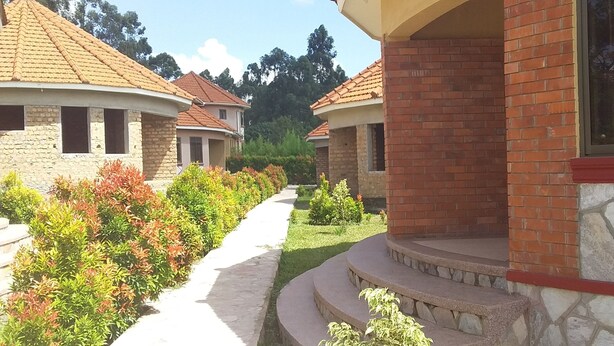 A residential area with houses featuring red-tiled roofs and brick exteriors. A pathway runs through the greenery, leading to a house with a stone staircase. - Mango Cottages & Recreation Center (Kakiri)