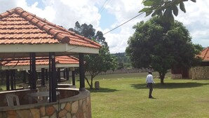 A covered outdoor seating area with stone walls and a tiled roof, surrounded by a grassy lawn and trees. - Mango Cottages & Recreation Center (Kakiri)