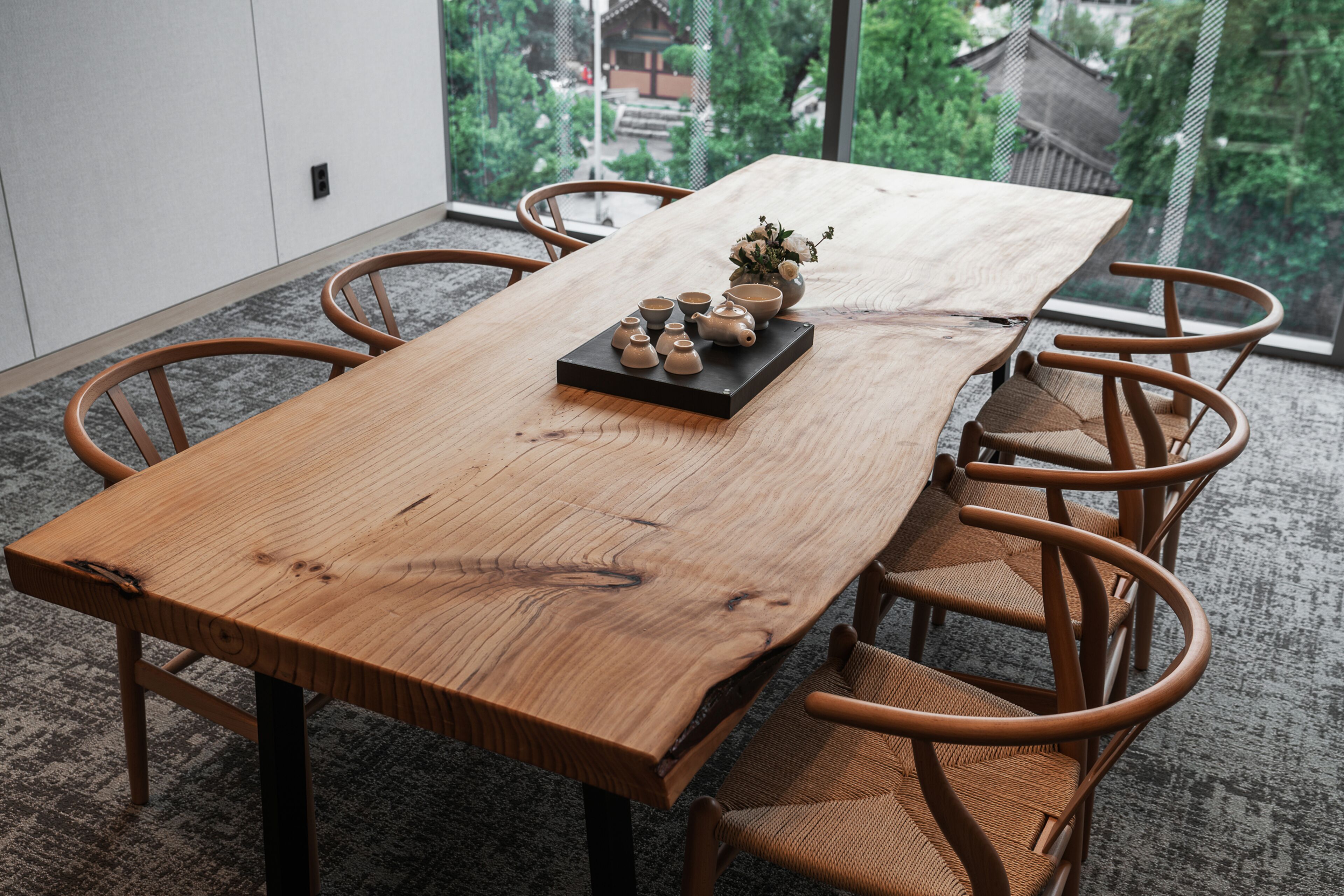 A wooden dining table with six wicker chairs, a black tray with ceramic teapots and cups, and a vase with flowers.