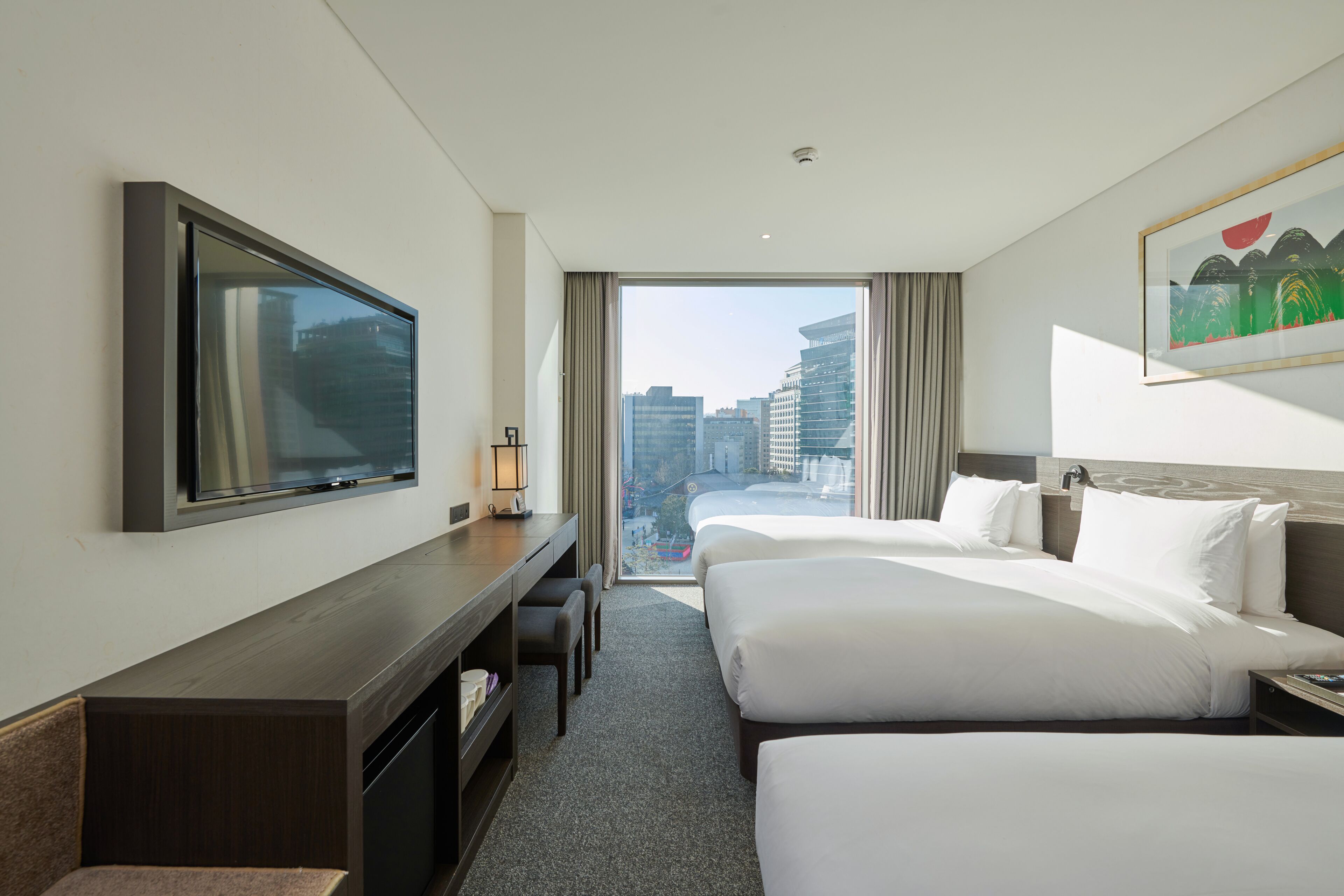 A hotel room with two beds, a TV, a desk, and a large window offering a city view.