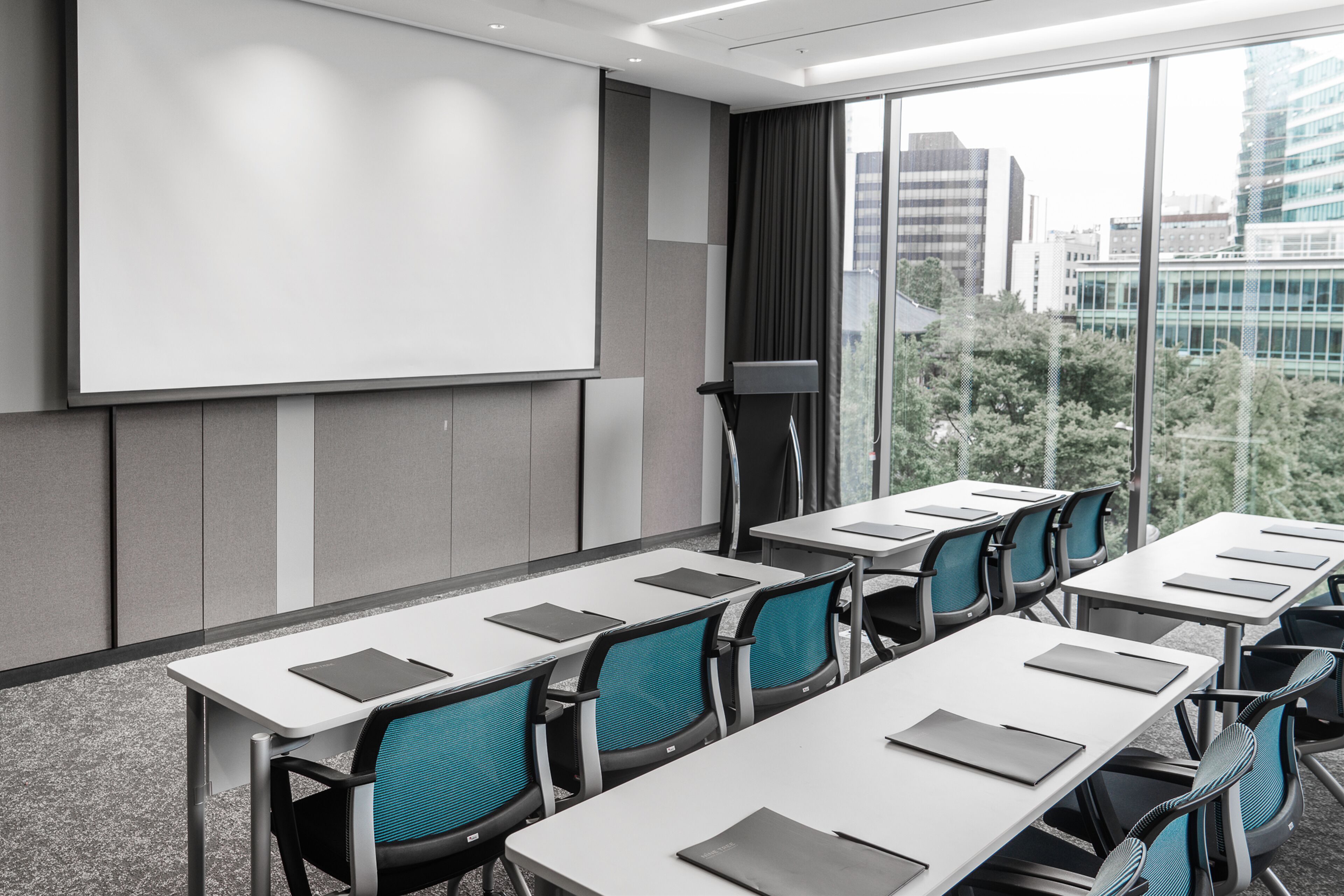 Ein Konferenzraum mit einer großen Projektionsleinwand, mehreren Tischen mit Stühlen und Blick auf die Stadt durch große Fenster.