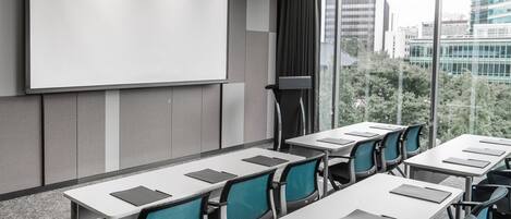 Un salón de conferencias con una pantalla de proyección grande, varias mesas con sillas y vistas a la ciudad a través de amplios ventanales.