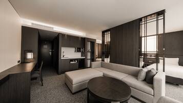 A modern hotel room with a sofa, a round coffee table, a desk, and a kitchenette.