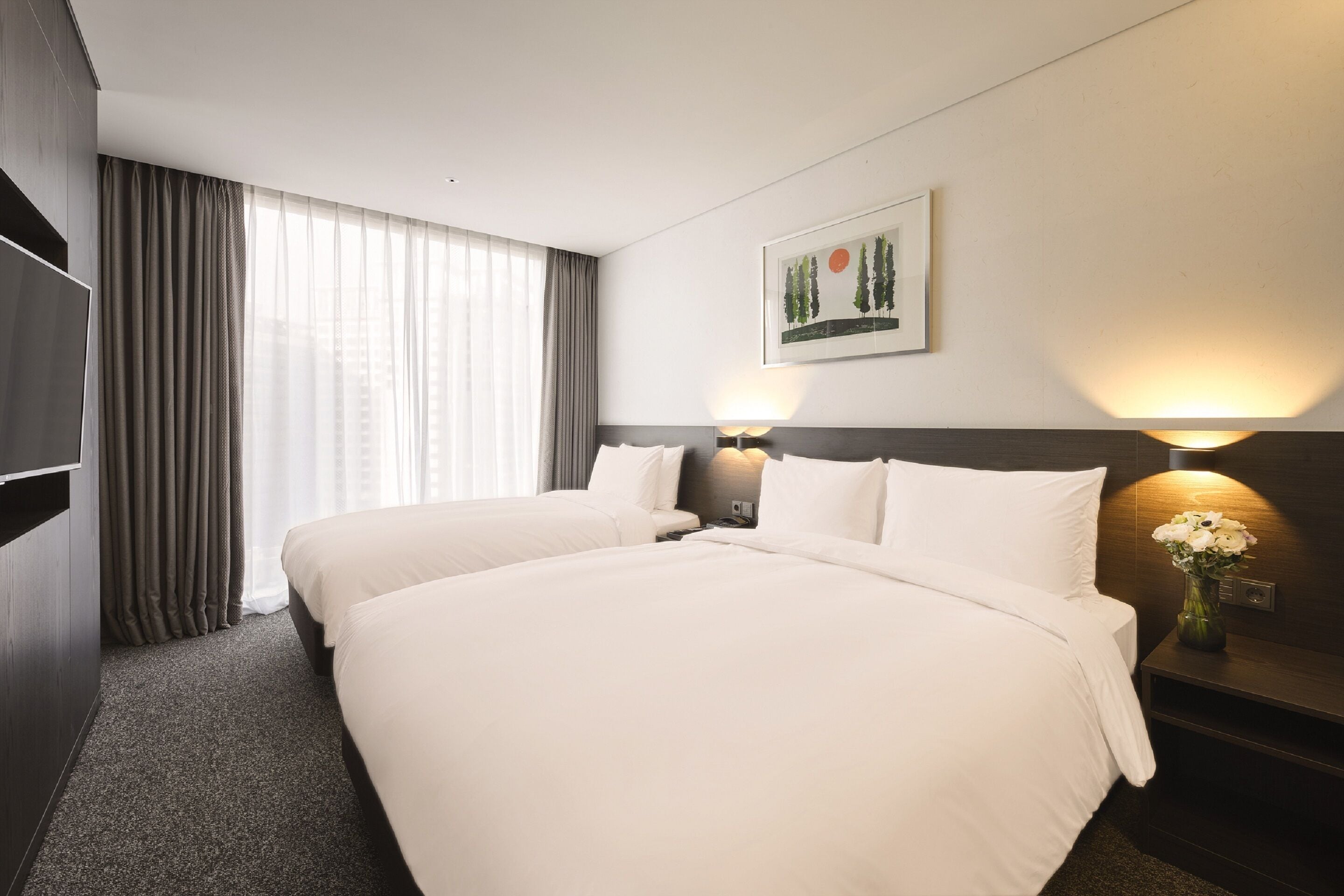 A hotel room with two beds, a large window with curtains, a painting on the wall, and a nightstand with a lamp and flowers.
