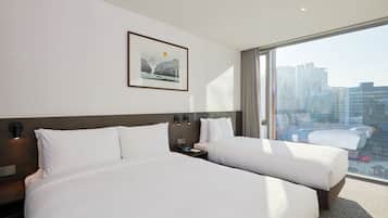 A hotel room with two beds, a nightstand, a wall-mounted lamp, a framed picture, and a large window with a city view.