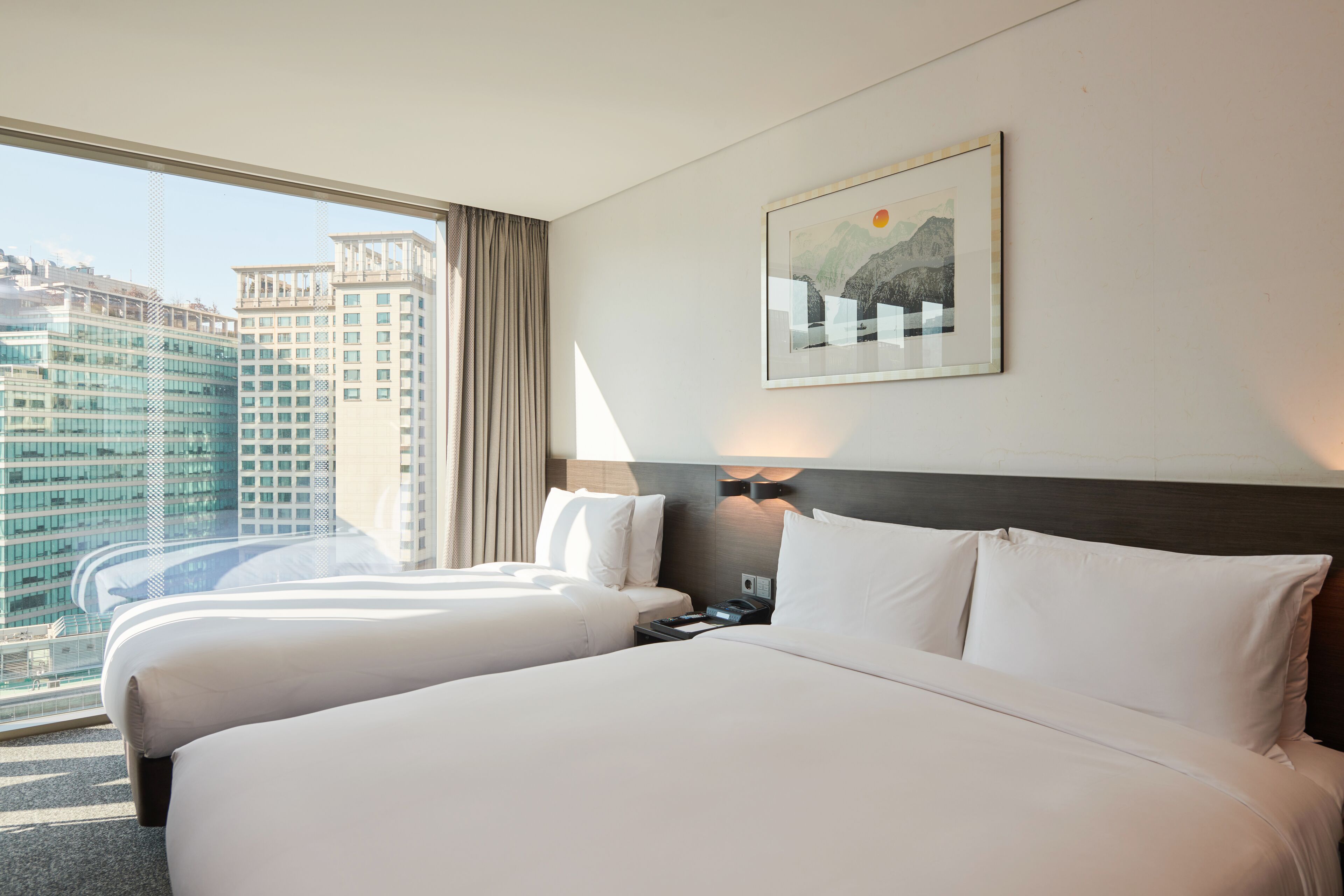 Habitación de hotel con dos camas, un amplio ventanal con vistas a la ciudad, una obra de arte enmarcada en la pared y una mesita de noche con lámpara.