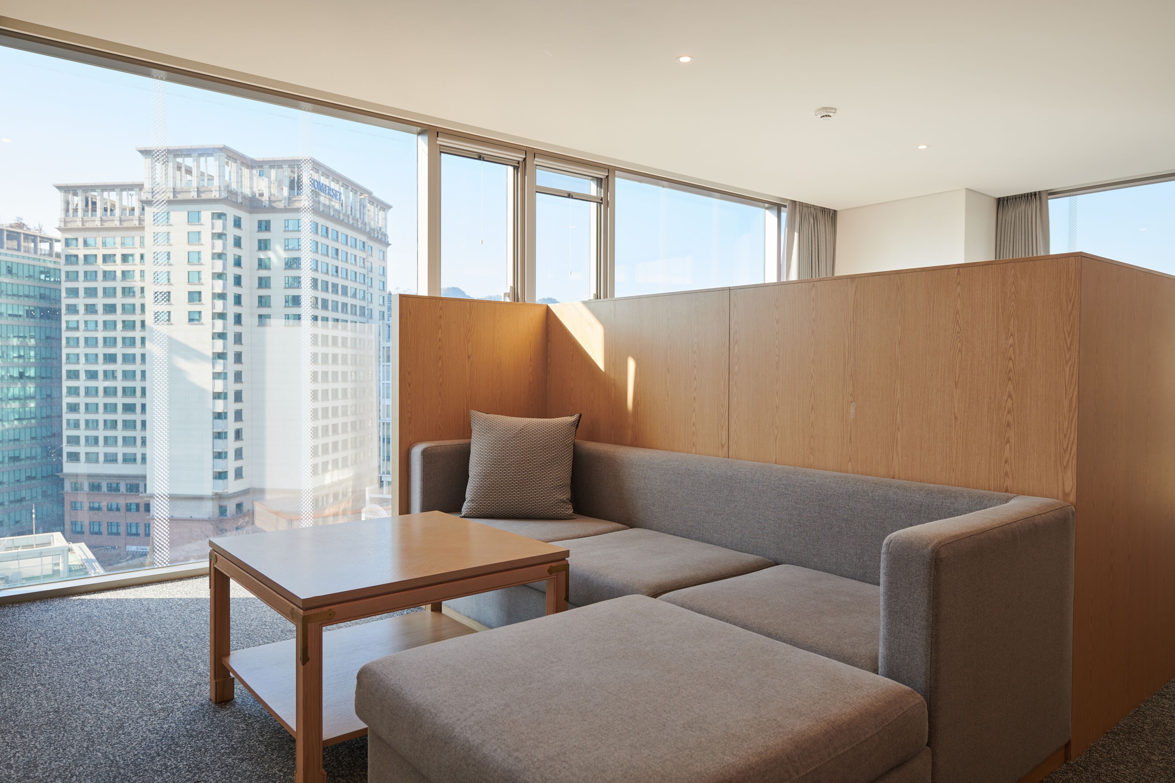 A modern hotel room with a grey sofa, a wooden table, and a large window offering a city view.