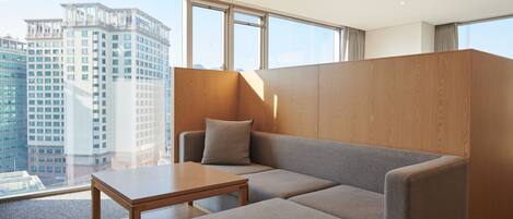 A modern hotel room with a grey sofa, a wooden table, and a large window offering a city view.