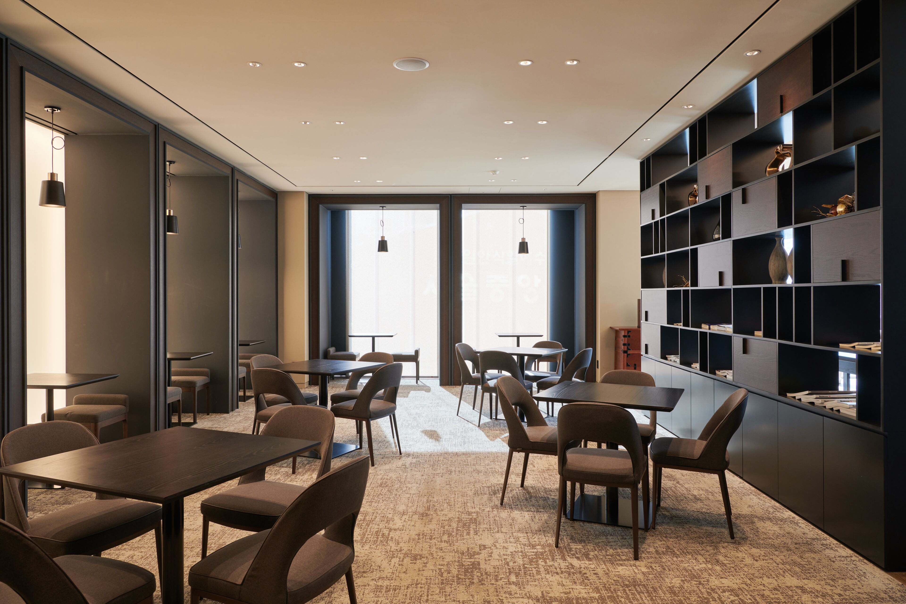 A modern dining area with tables and chairs, large windows, and a bookshelf with decorative items.
