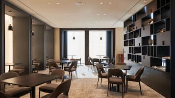 A modern dining area with tables and chairs, large windows, and a bookshelf with decorative items.
