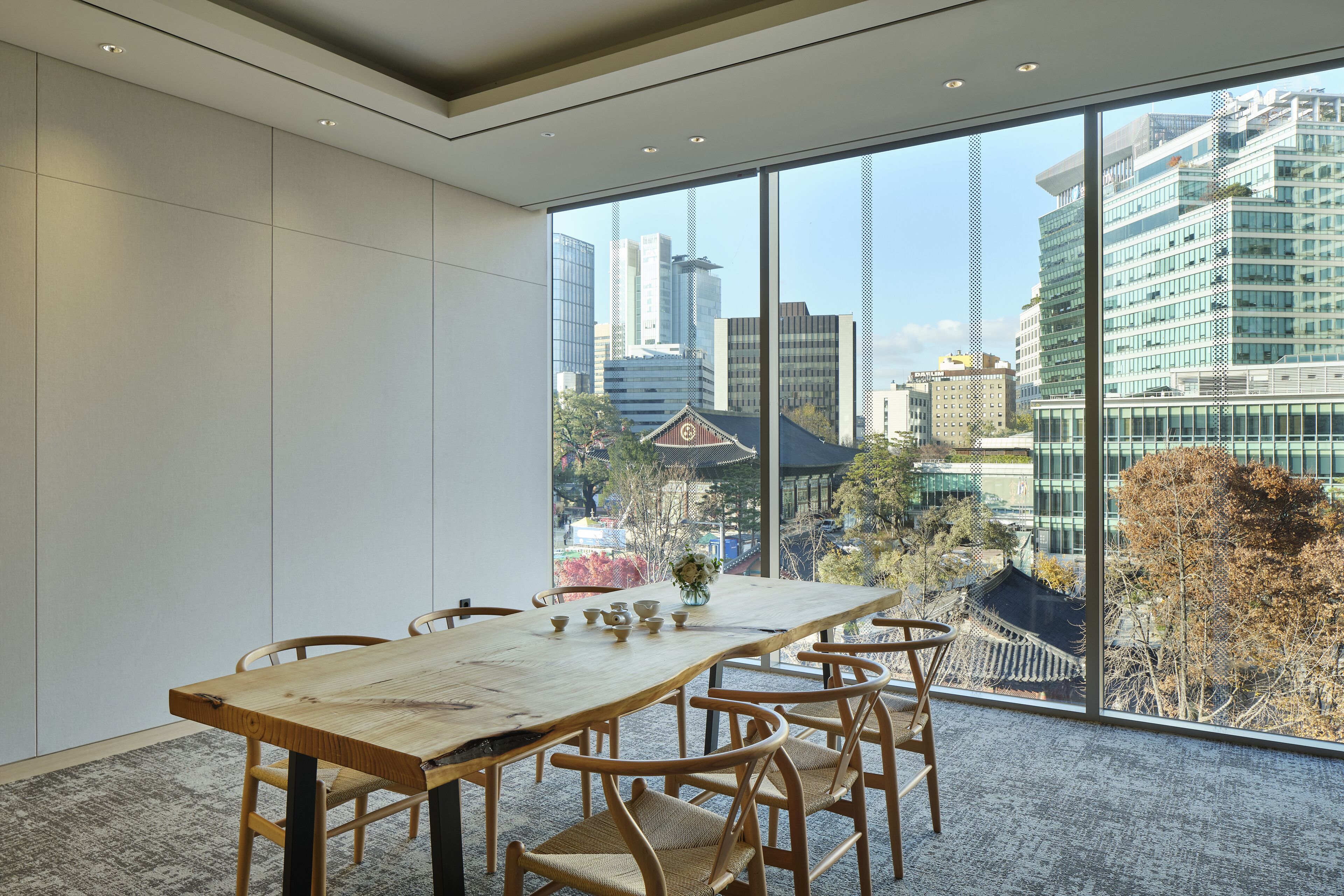 Un comedor moderno con una larga mesa de madera y sillas, ventanas amplias que ofrecen vistas de edificios de la ciudad y arquitectura tradicional.