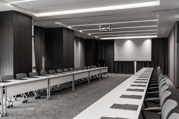 Un salón de conferencias con una larga mesa, sillas, una pantalla de proyector y suelo alfombrado.