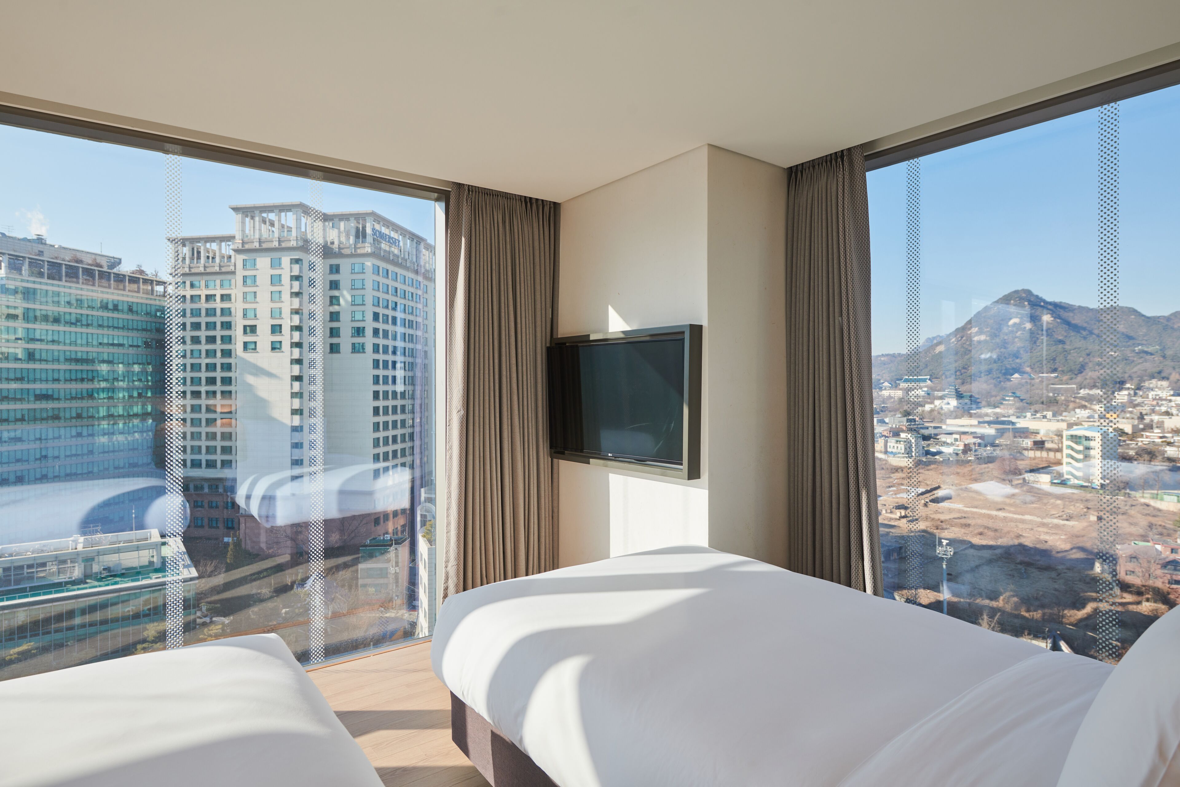 A hotel room with a large bed, a TV mounted on the wall, and a city view through large windows.
