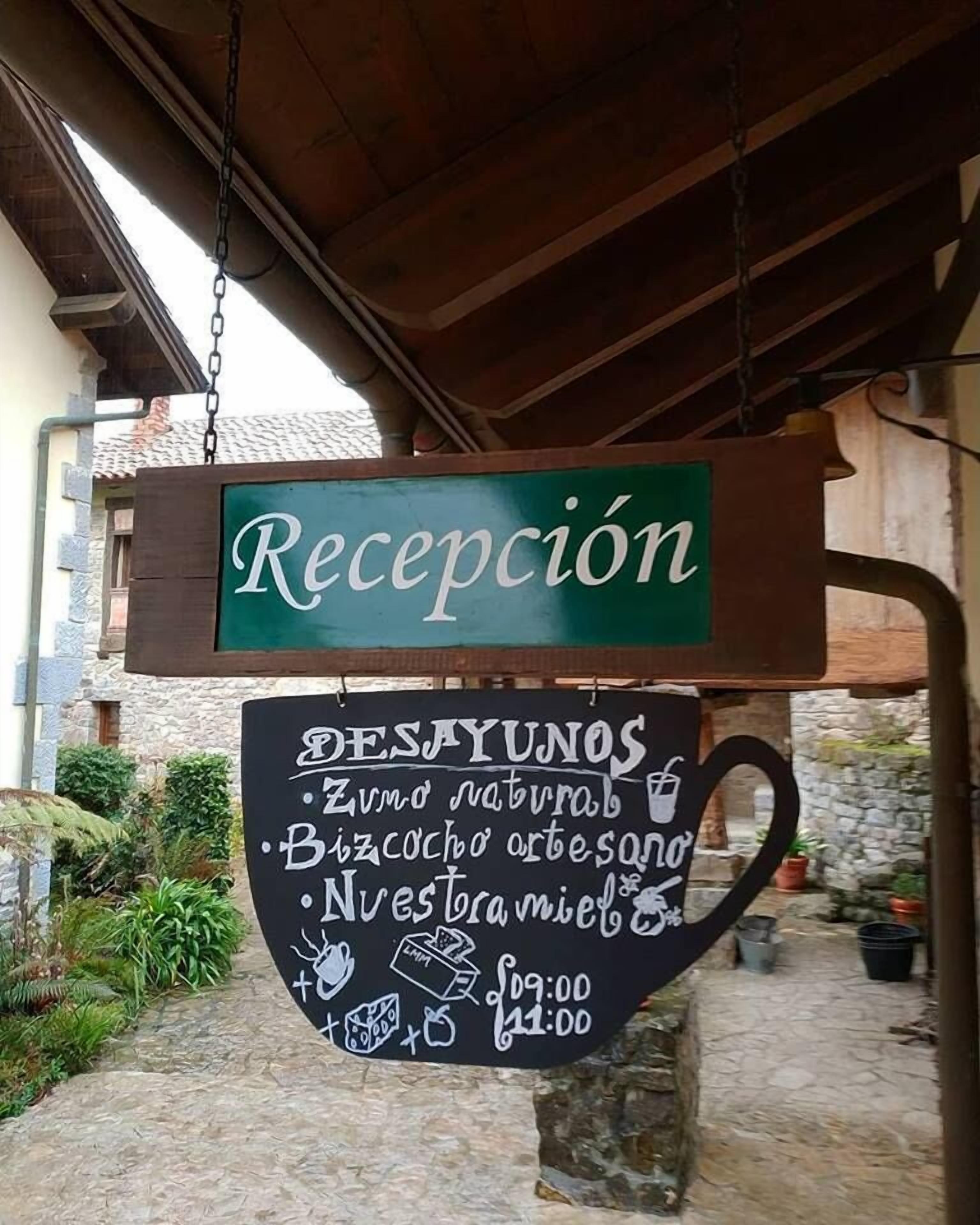Une enseigne d’accueil avec un fond vert et du texte blanc, suspendue à une structure en bois. En dessous, un tableau noir affiche les différentes options et les prix du petit-déjeuner.