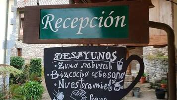 Une enseigne d’accueil avec un fond vert et du texte blanc, suspendue à une structure en bois. En dessous, un tableau noir affiche les différentes options et les prix du petit-déjeuner.