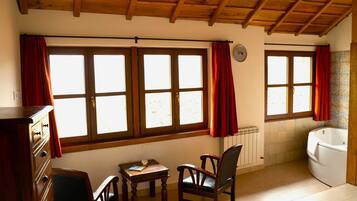 Una habitación con vigas de madera en el techo, un aparador de madera, una mesita con un libro y un vaso, dos sillas de madera, una bañera blanca y ventanas amplias con cortinas rojas.