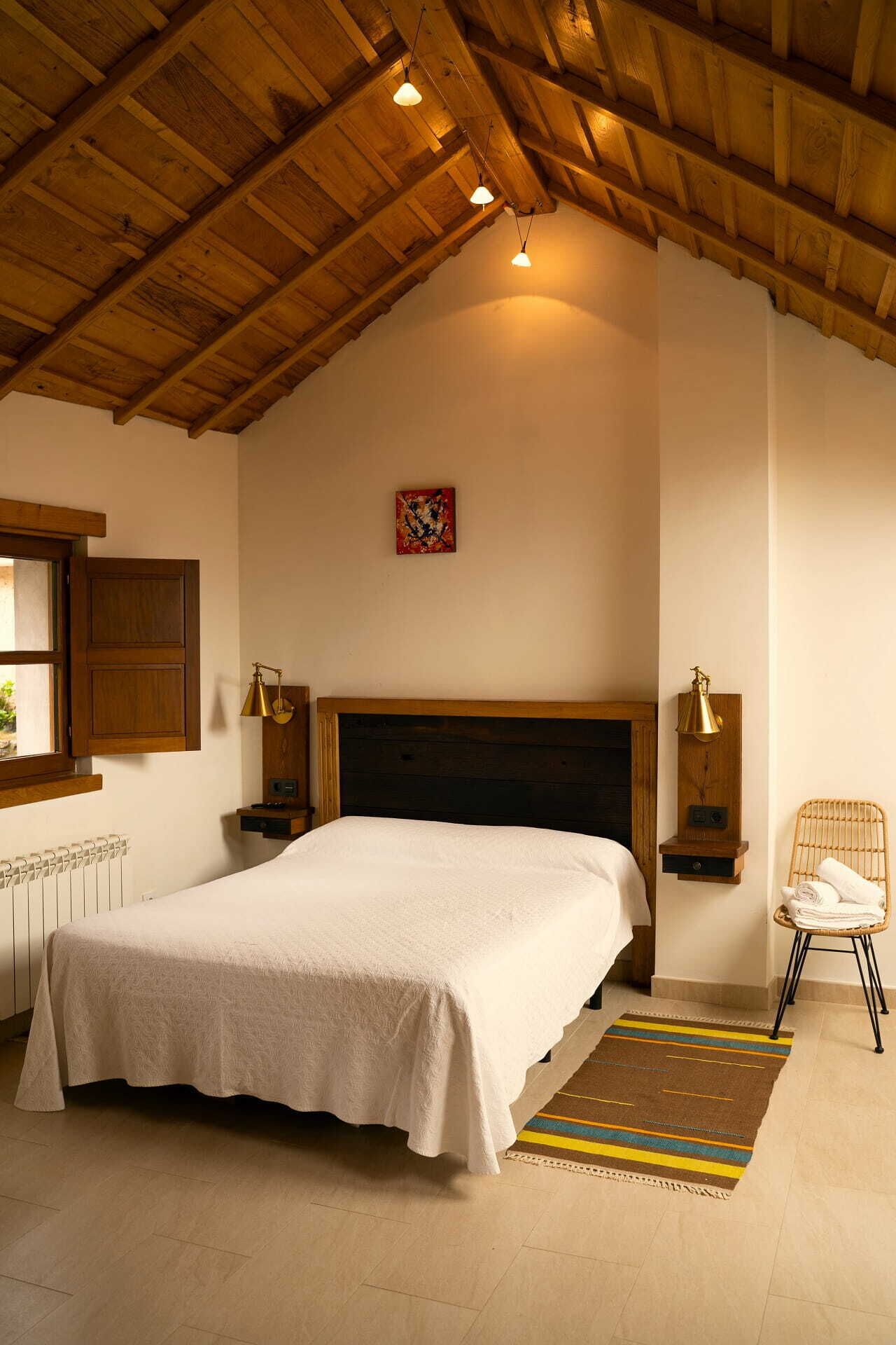 A bedroom with a wooden ceiling, a bed with a white bedspread, a nightstand with a lamp, a chair, and a small rug.