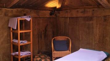 A wooden room with a massage table, a wooden shelf with towels, and a wicker chair.