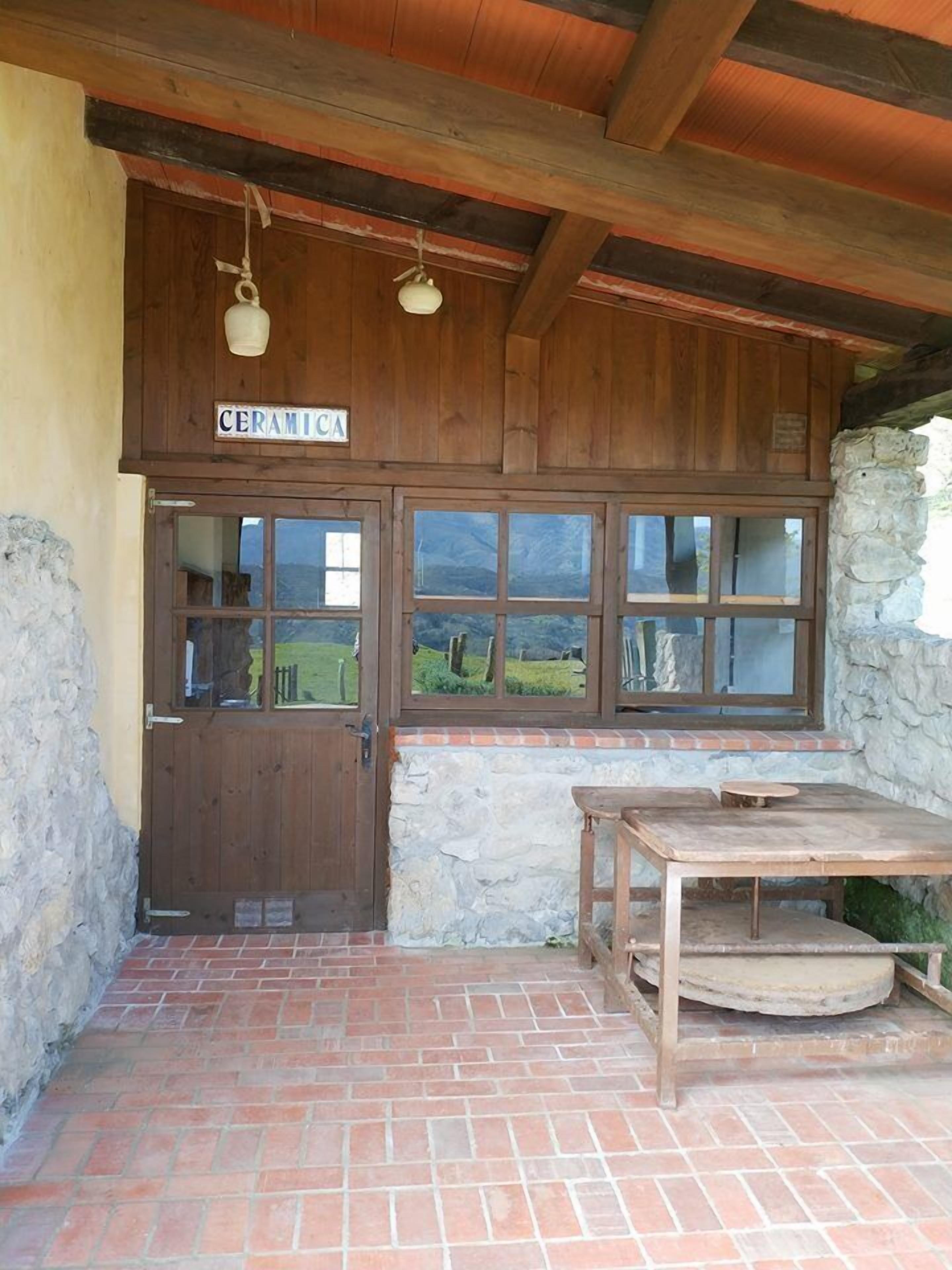 A rustic wooden building with a sign reading "CERAMICA." It features a wooden door, a stone wall, and a brick-paved patio with a wooden table and bench.