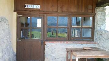 A rustic wooden building with a sign reading "CERAMICA." It features a wooden door, a stone wall, and a brick-paved patio with a wooden table and bench.