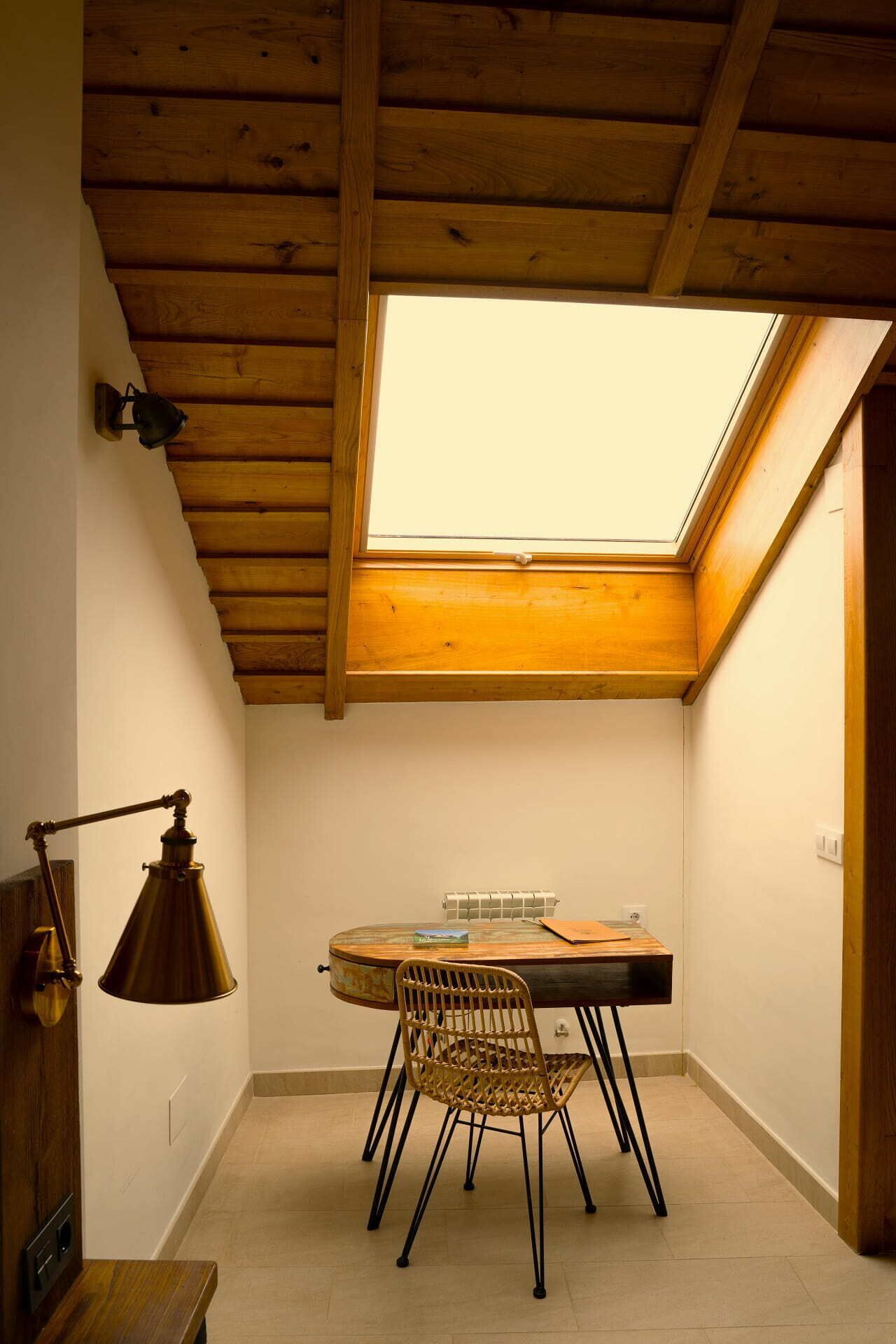 Una habitación pequeña y acogedora con tragaluz, una mesa redonda de madera, una silla de mimbre y una lámpara antigua.