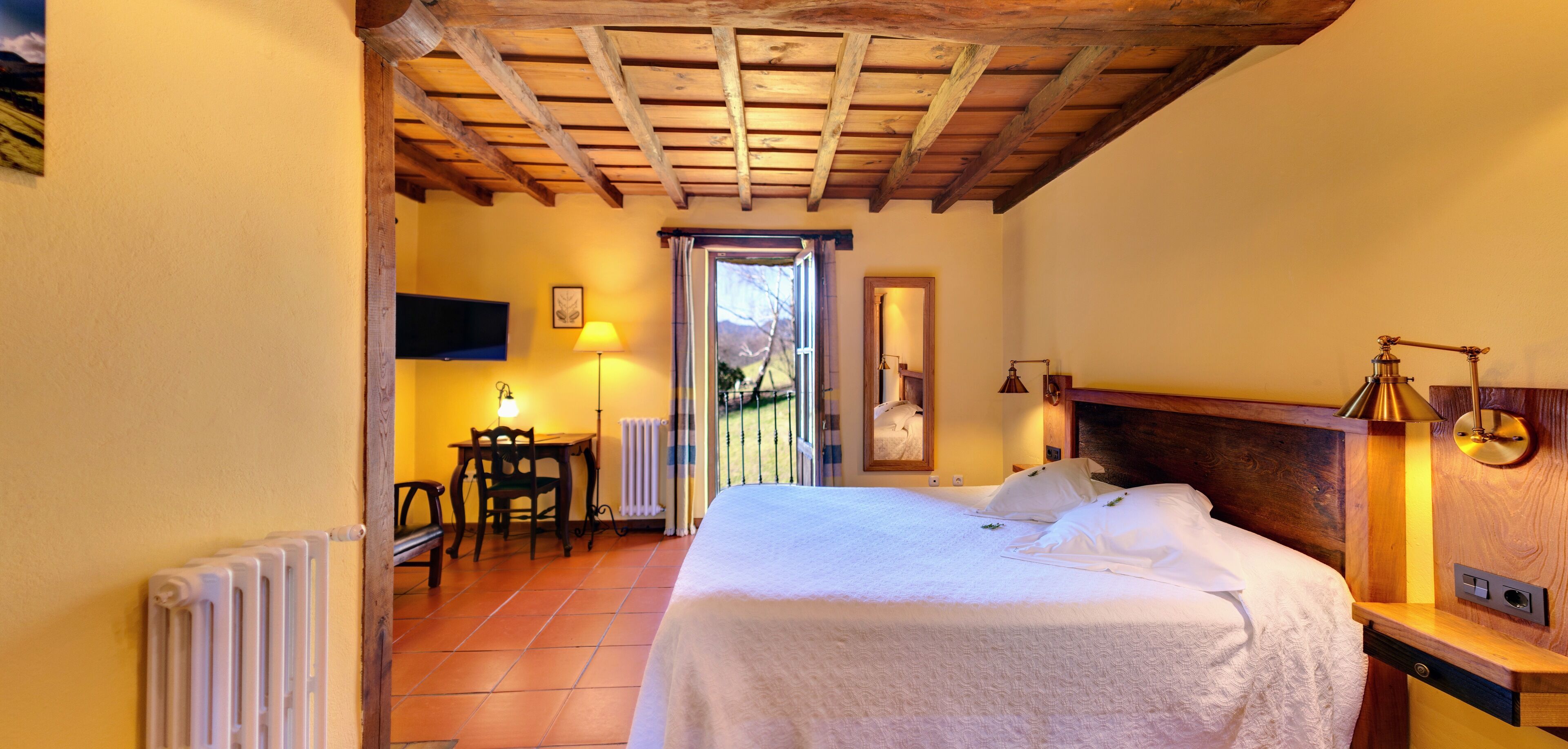 A bedroom with a wooden ceiling, a bed with white linens, a bedside table with a lamp, and a view through a door to an outdoor area.