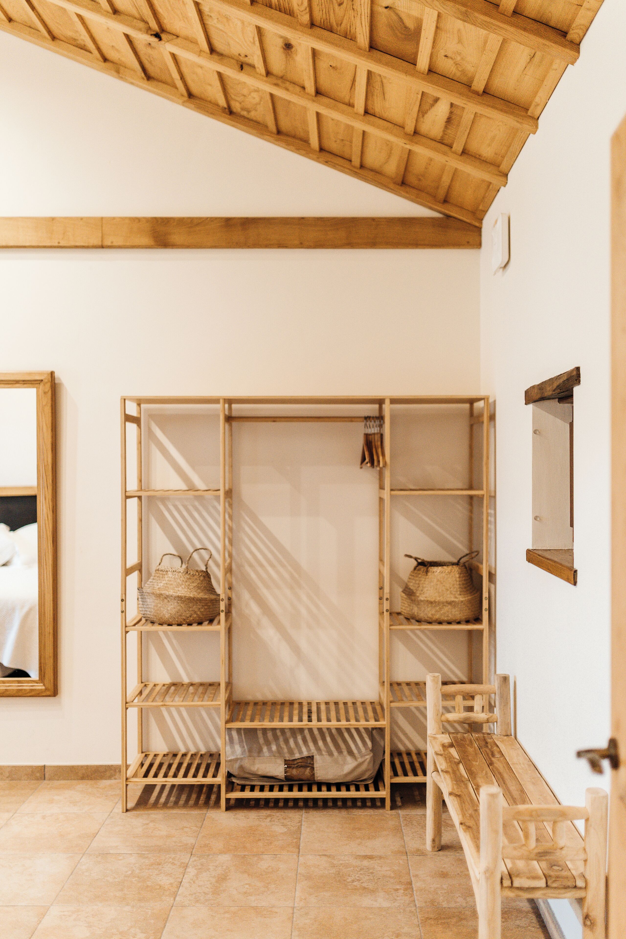 Un banco de madera, un armario de madera con estantes y un espejo en una pared blanca.