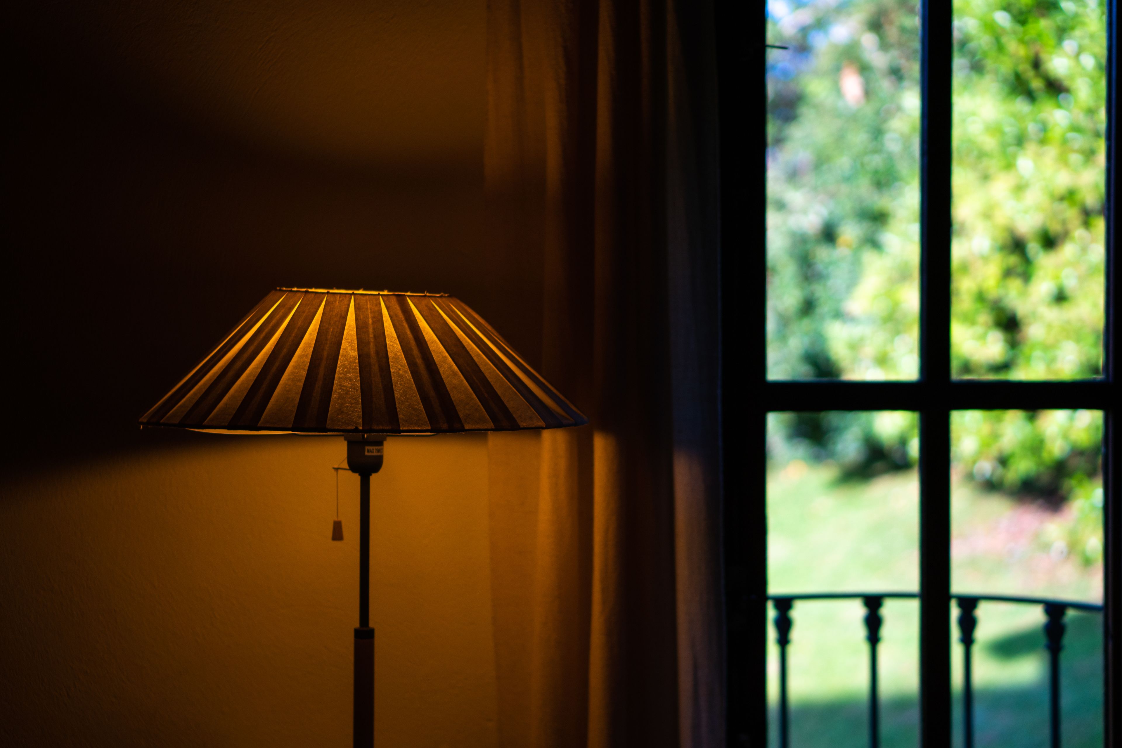 A lamp with a striped shade next to a window with a view of greenery.