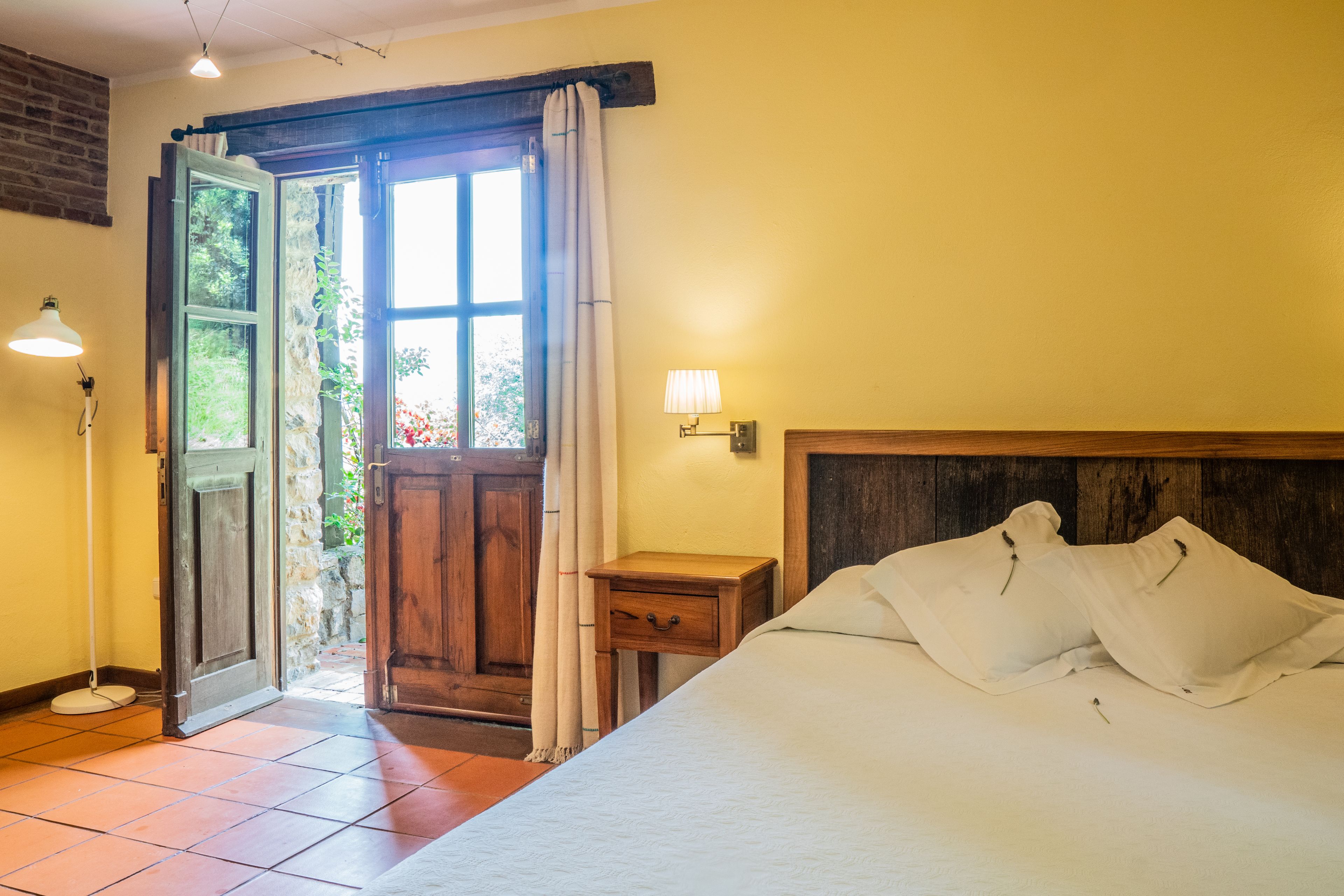 Ein Schlafzimmer mit Bett, Nachttisch, Lampe, Holztür und Blick ins Freie.