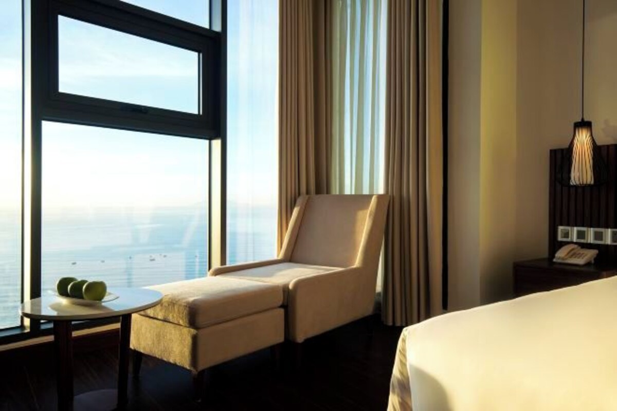 A hotel room with a large window, a bed, a chair, a small table with apples, and a hanging light fixture.
