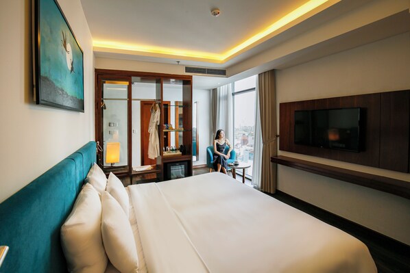 A hotel room with a large bed, a TV mounted on the wall, a desk with a chair, and a view of the city through large windows. - Maximilan DaNang Beach Hotel (Da Nang)