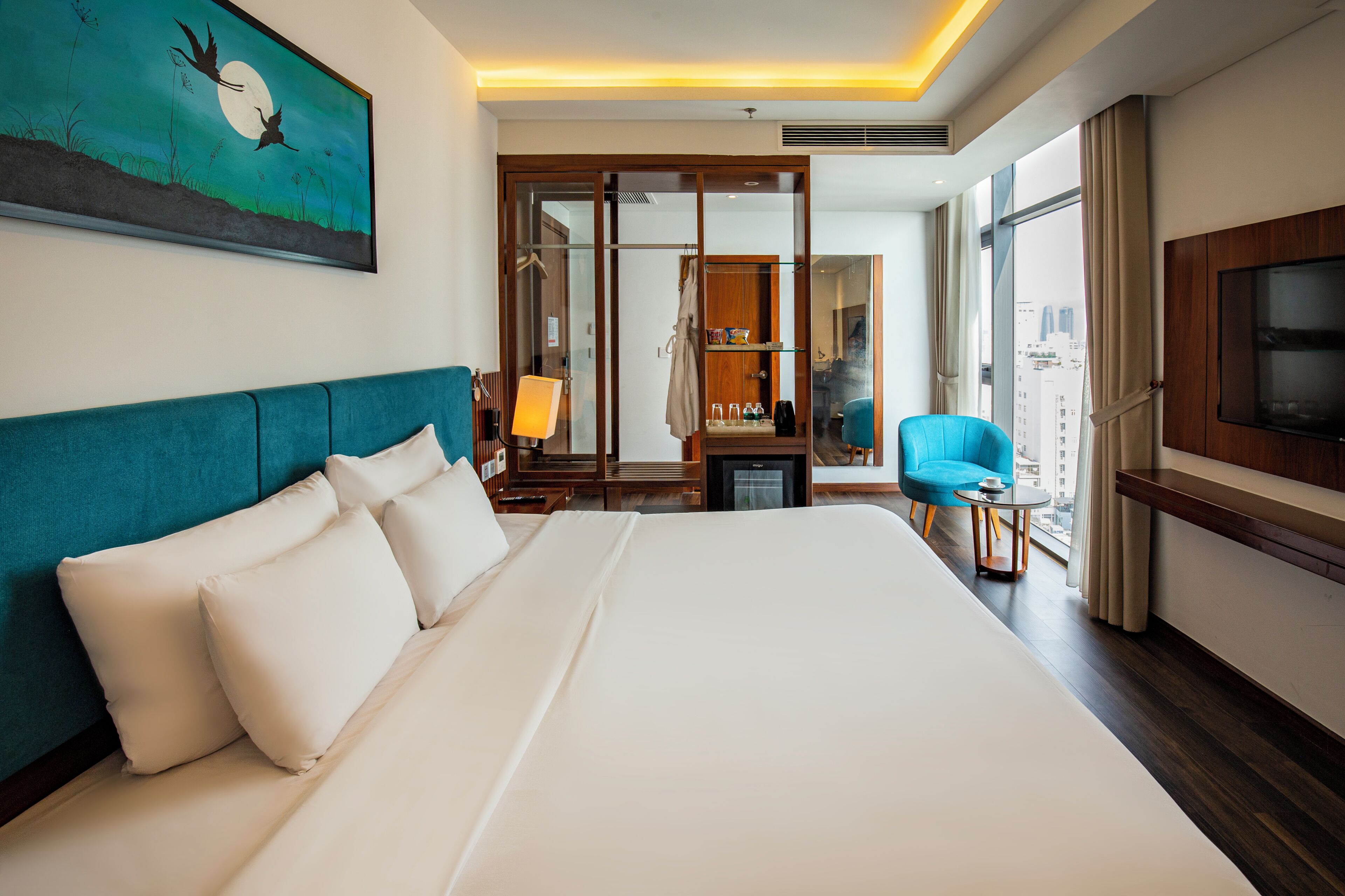 A hotel room with a large bed, a TV, a desk with a chair, a wardrobe, and a view of the city.