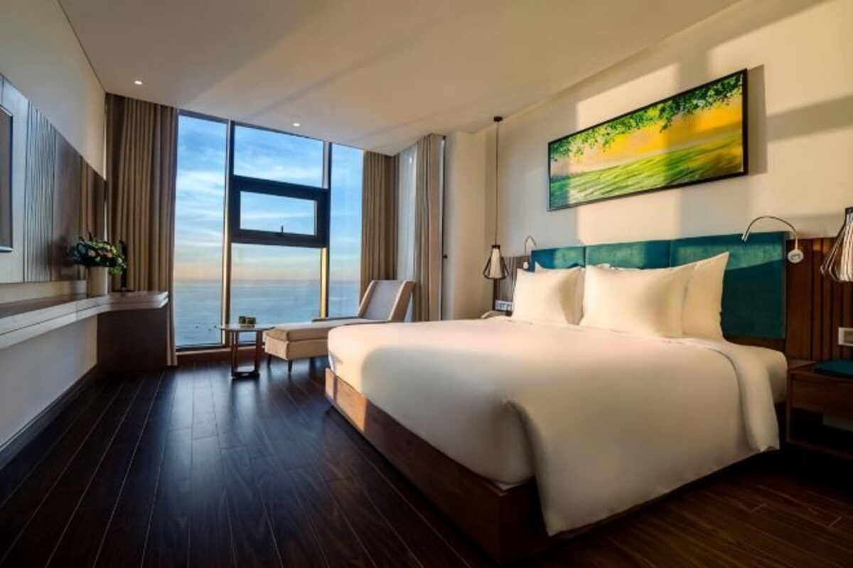 A hotel room with a large bed, a view of the sea, a painting on the wall, and a desk with a chair.